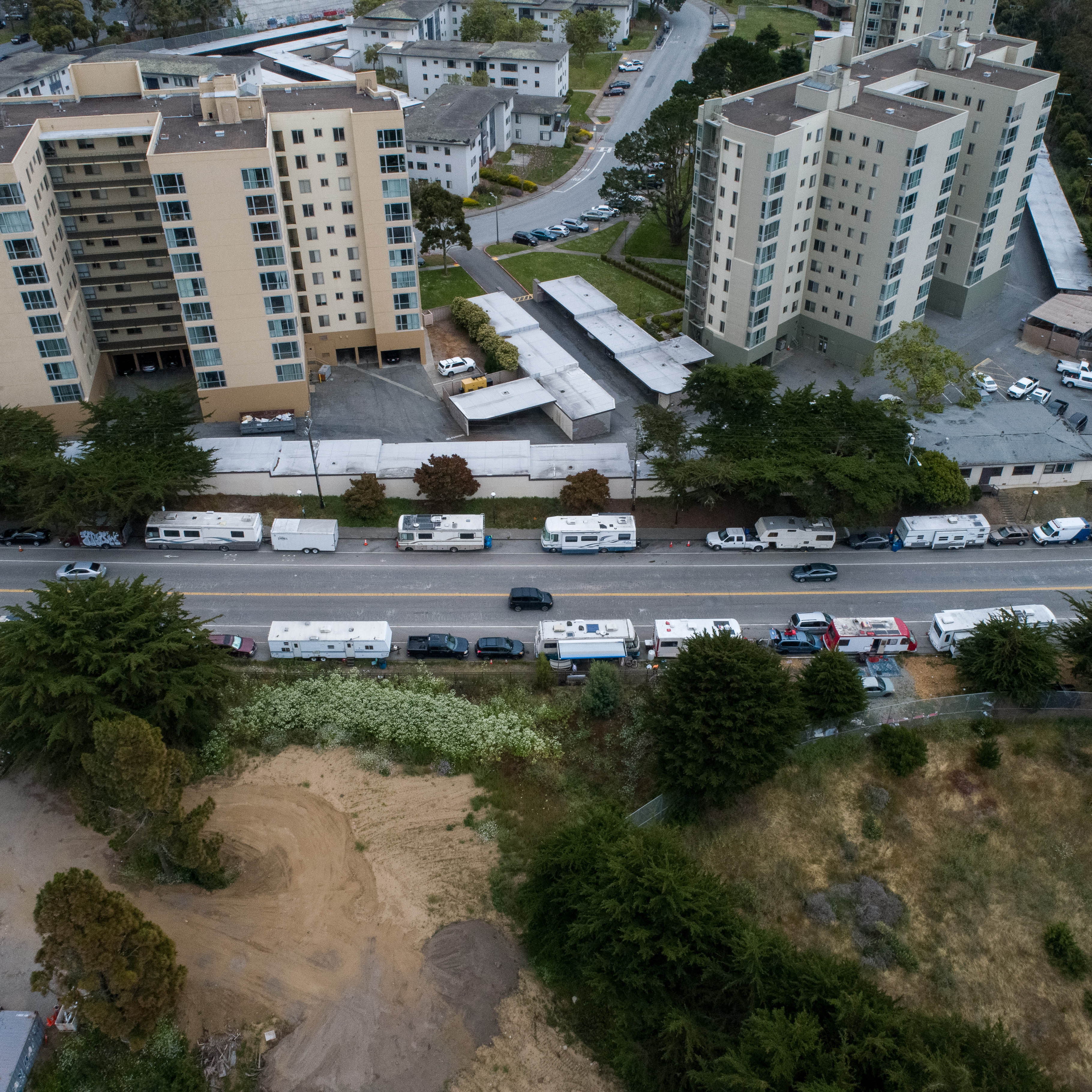 The image shows a row of recreational vehicles (RVs) parked along a street beside a series of high-rise apartment buildings and a green, grassy area with trees.