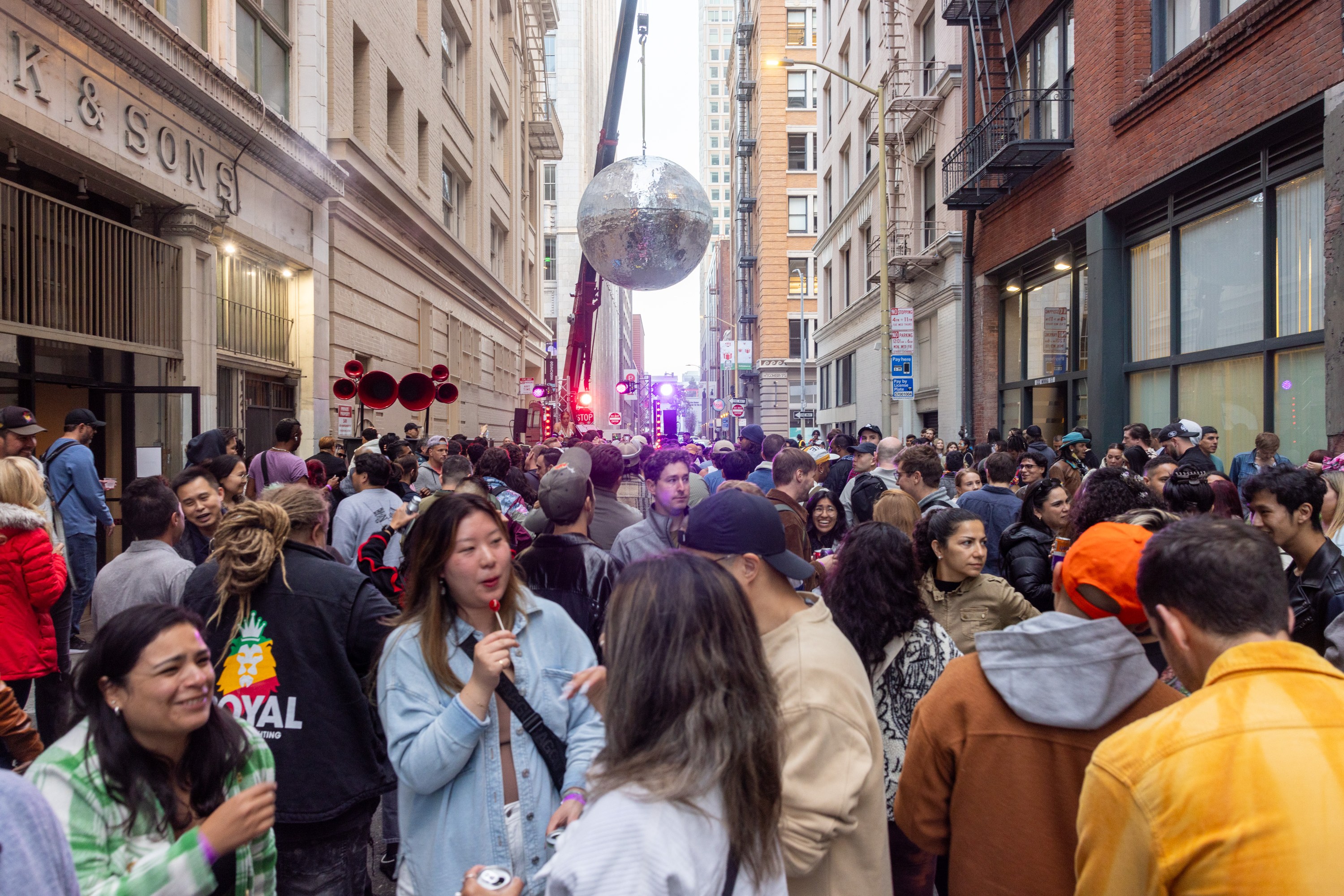 A crowded street party in a city with people mingling, eating, and talking. Large silver disco ball hanging mid-street. Buildings line both sides, creating an alley-like effect.