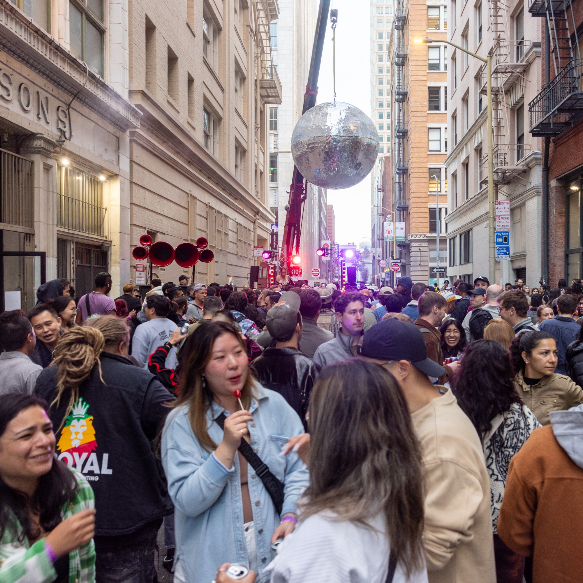 A crowded street party in a city with people mingling, eating, and talking. Large silver disco ball hanging mid-street. Buildings line both sides, creating an alley-like effect.