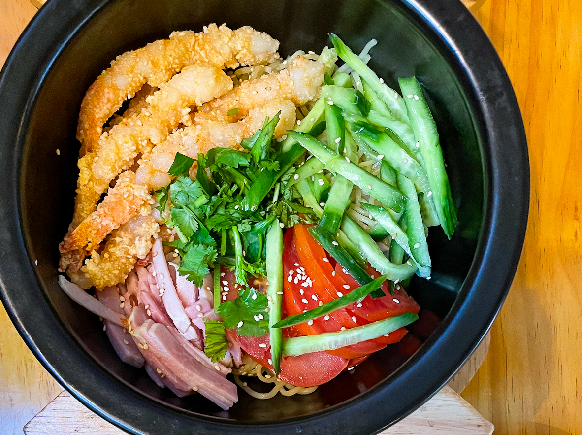 This image shows a bowl of food containing crispy fried strips, sliced cucumber, julienned ham, tomatoes, and cilantro, topped with sesame seeds.