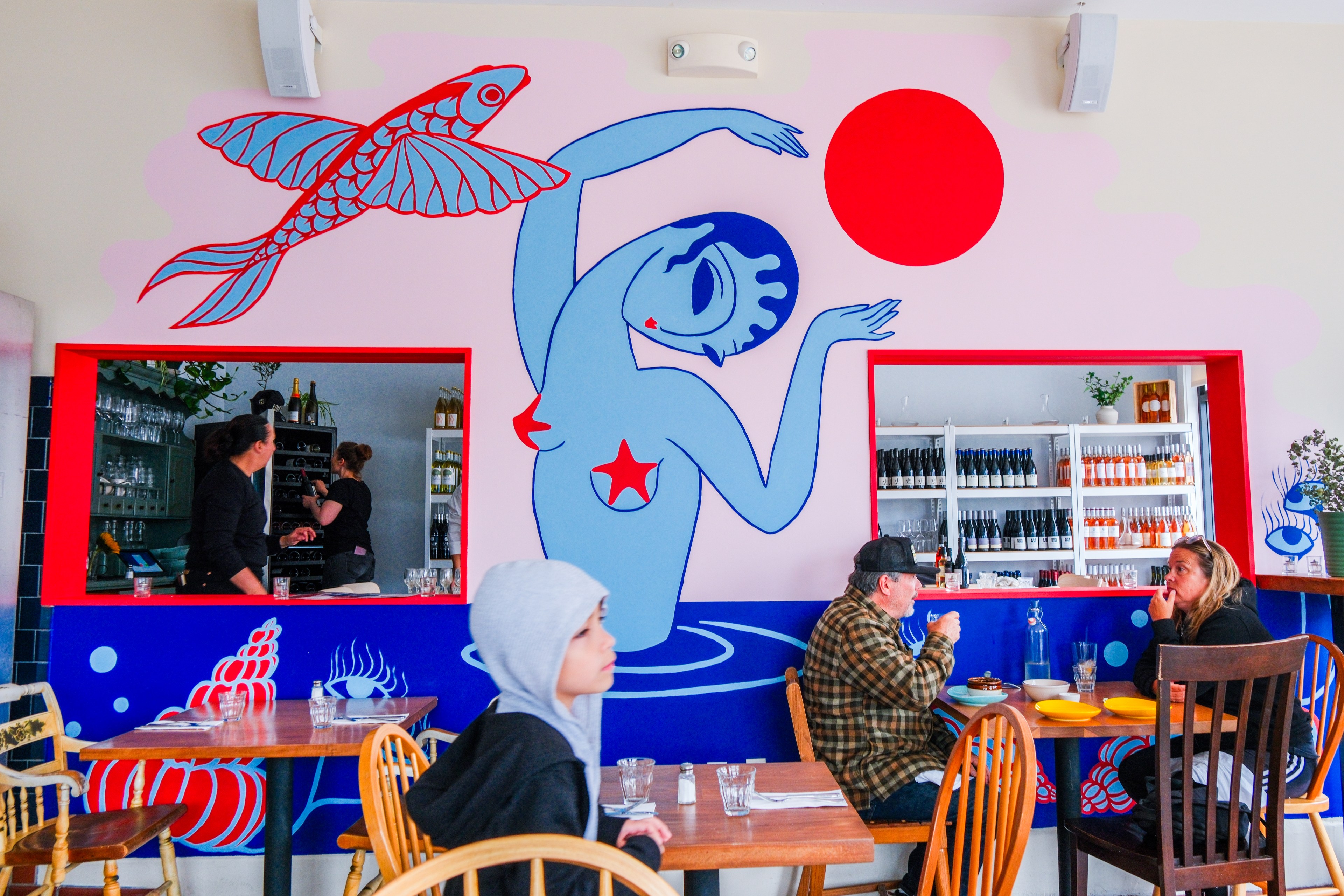 A colorful mural depicts a blue, stylized woman with a red ball and a flying fish. People dine at wooden tables, and two servers work near shelves of bottles.