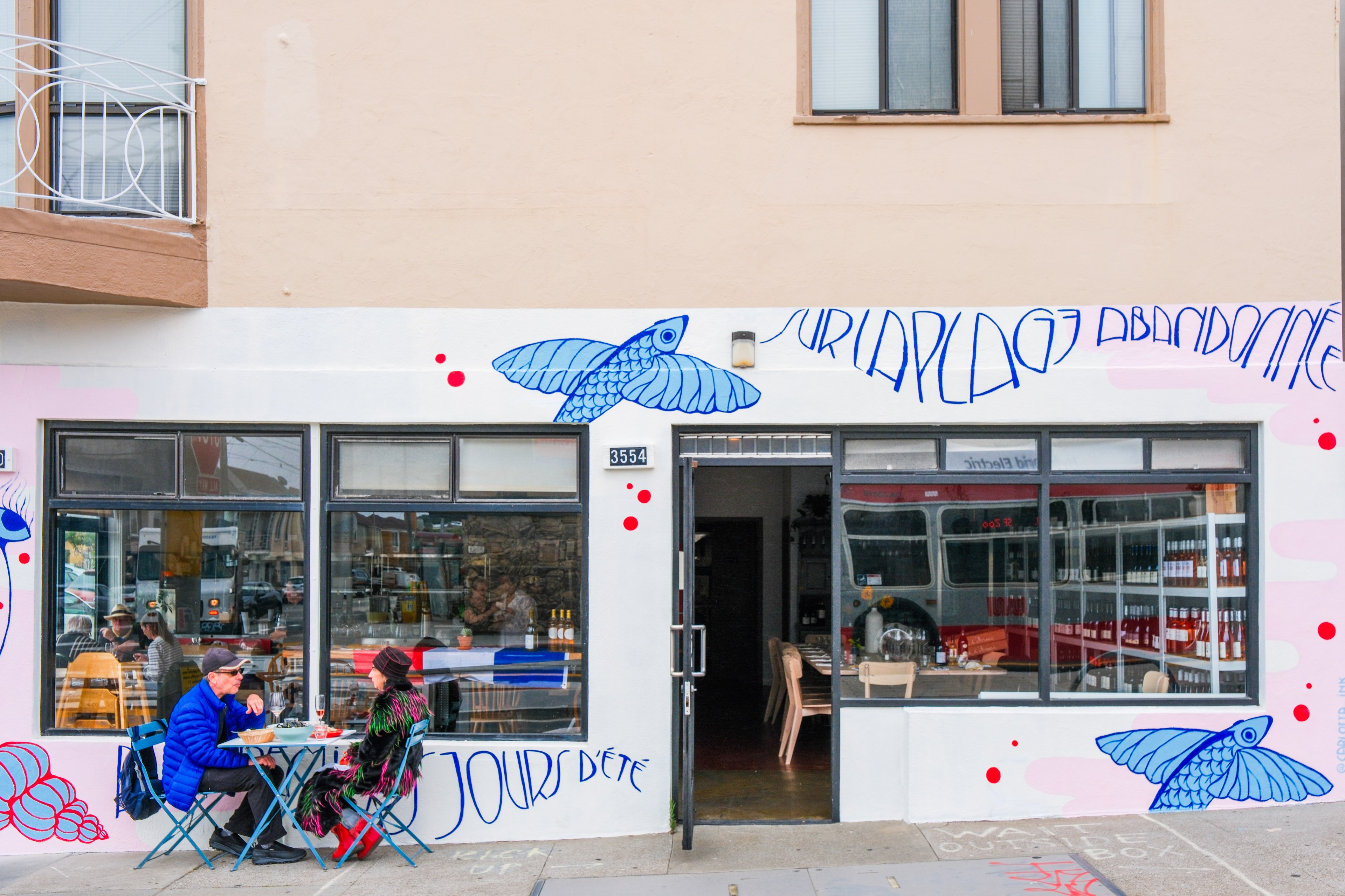 A small storefront cafe has blue mural art, featuring birds and French text, with two people dining at a blue table outside.