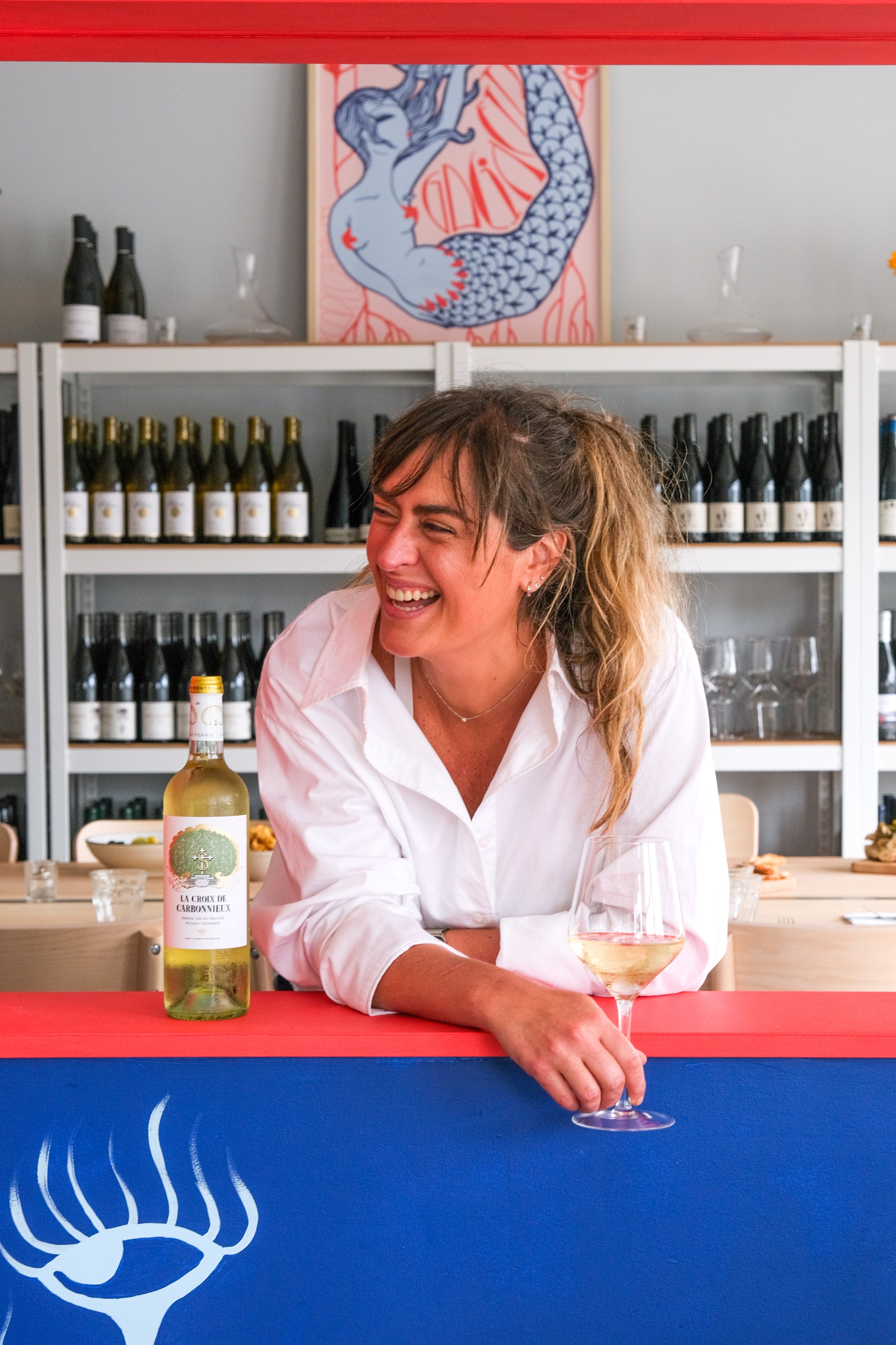 A smiling woman in a white shirt holds a glass of white wine, leaning on a counter with a blue and red backdrop. Shelves of wine bottles and a mermaid painting are behind her.