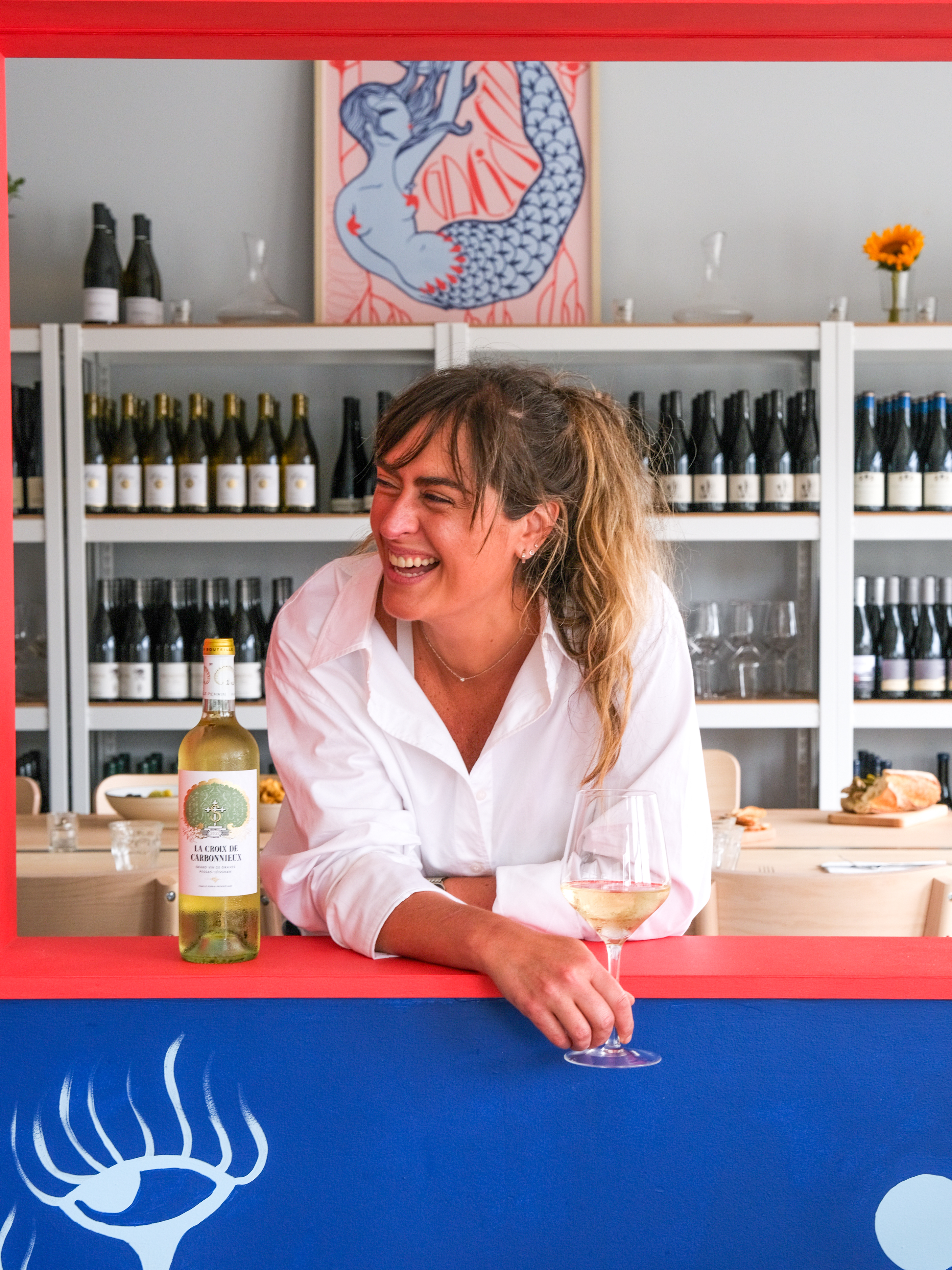 A smiling woman in a white shirt holds a glass of white wine, leaning on a counter with a blue and red backdrop. Shelves of wine bottles and a mermaid painting are behind her.