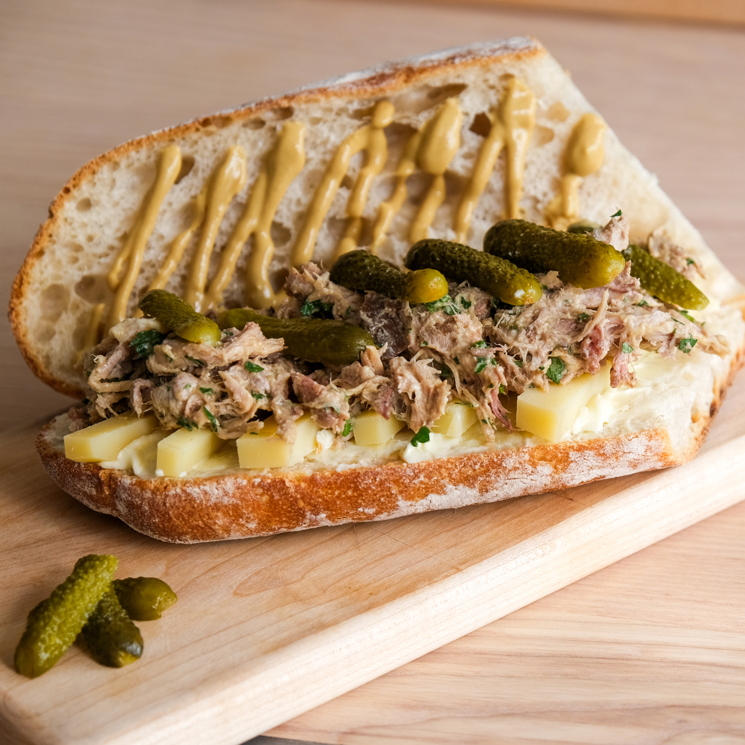 A sandwich on crusty bread with layers of sliced cheese, shredded meat, pickles, and mustard drizzled on the top half, served on a wooden board with extra pickles.