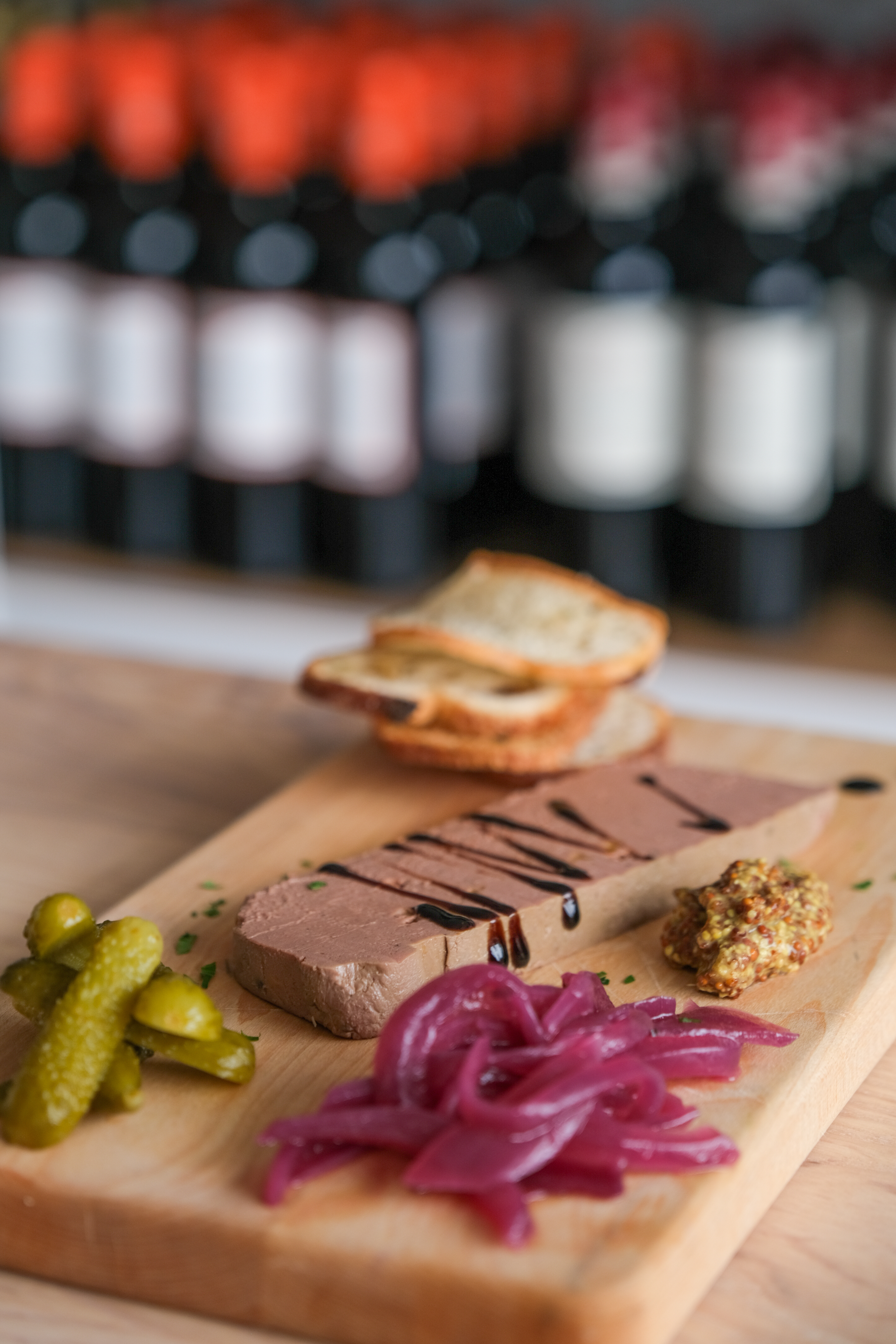 A wooden board holds a slice of pâté drizzled with a dark sauce, pickles, red onions, whole grain mustard, and toasted bread, against a blurred background of wine bottles.