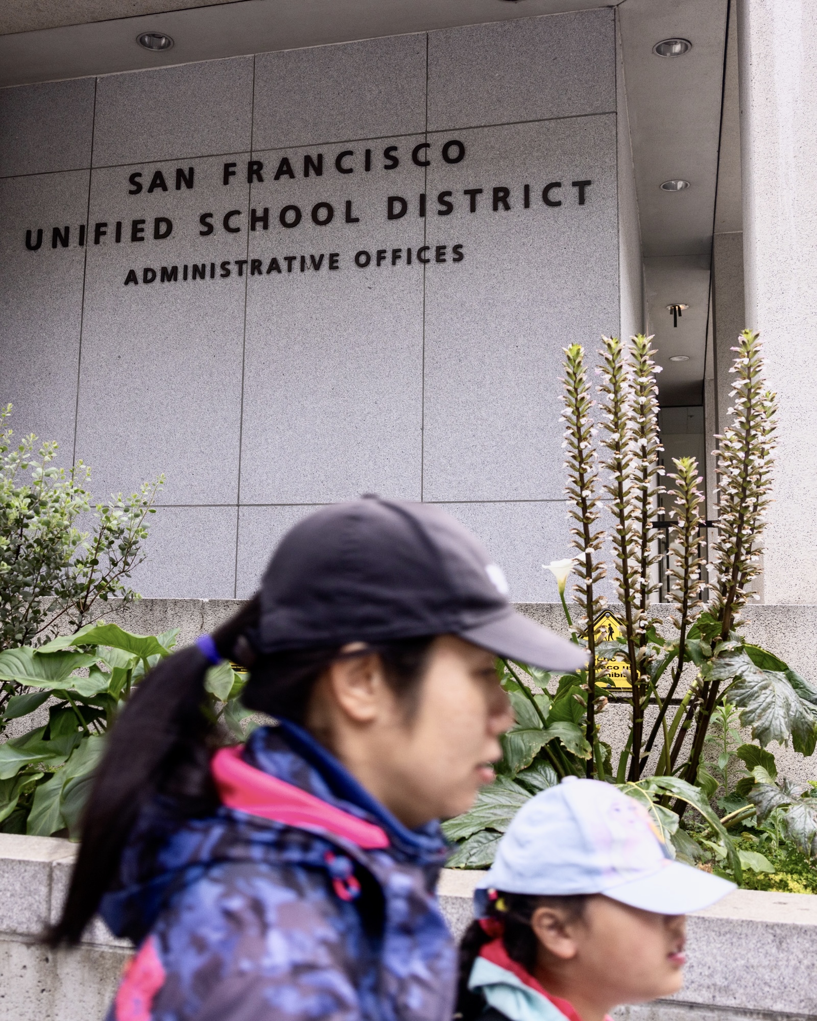 Two people walk past the San Francisco Unified School District Administrative Offices, with large plants in the foreground. The office name is displayed on the building.