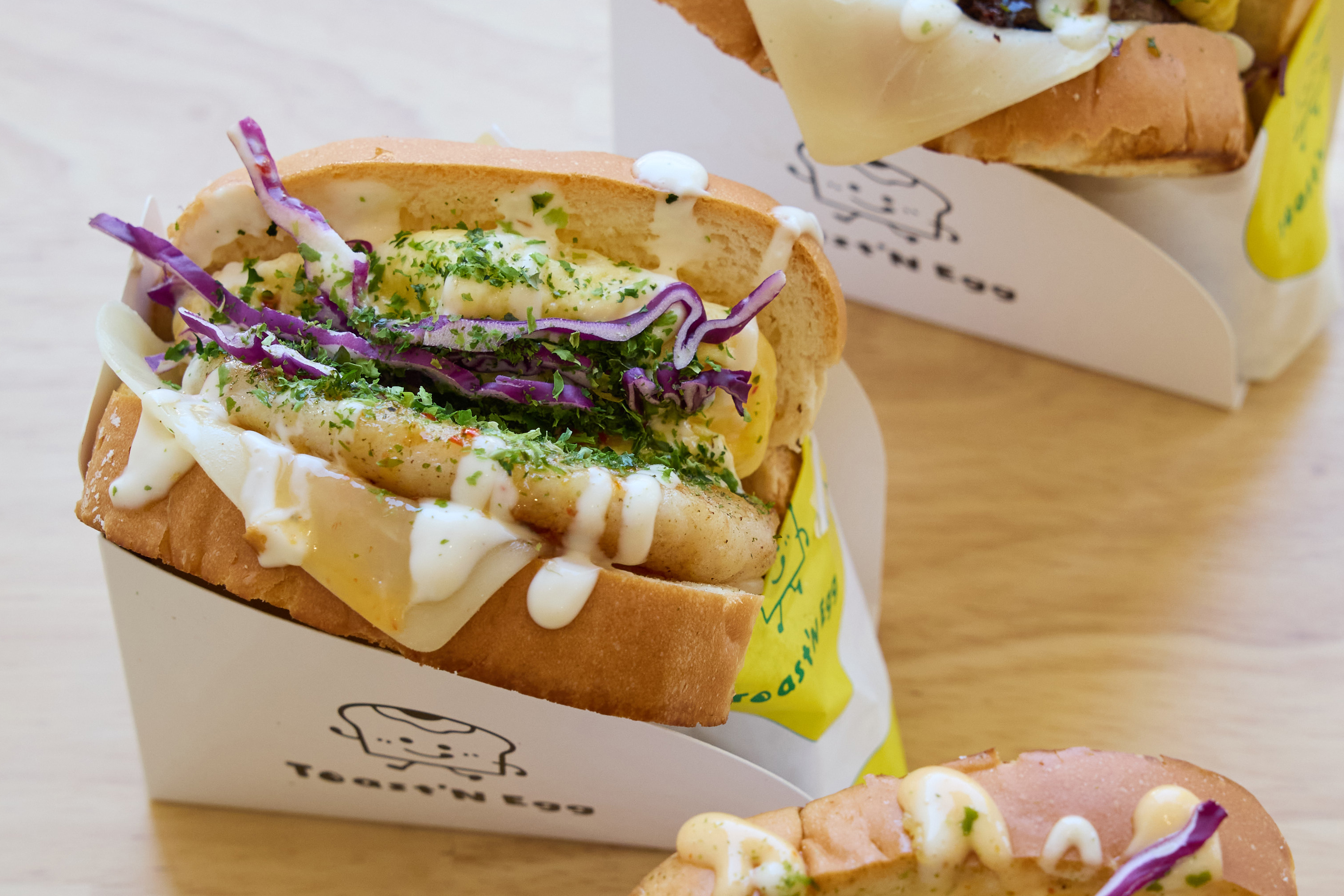 Three sandwiches in white containers have fluffy bread, topped with cheese, purple cabbage, sauces, and herbs. The label on the container shows a cute toast character.