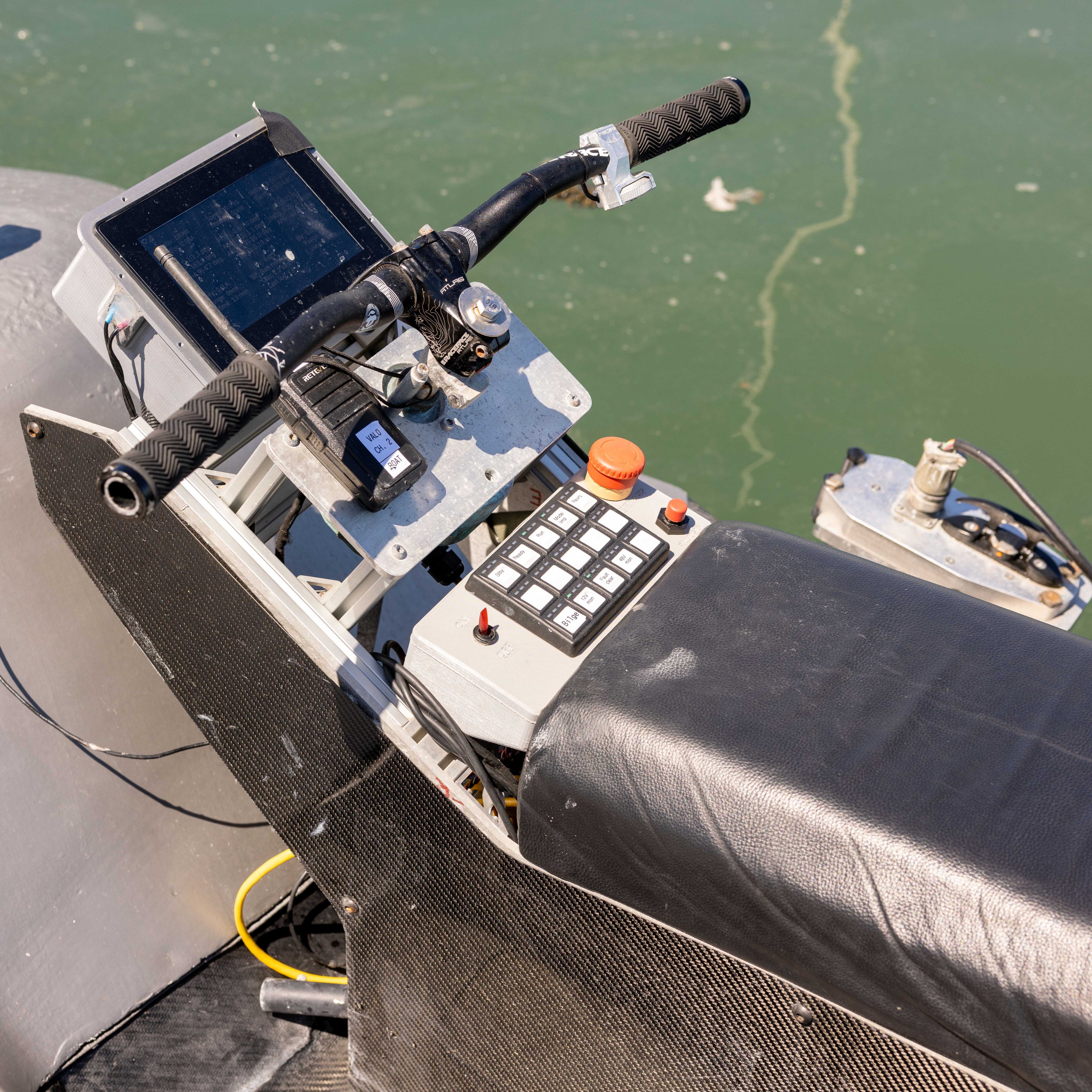 The image shows a boat's control console with a handlebar, a mounted tablet, buttons, switches, and a joystick on a sleek, cushioned seat by the water.