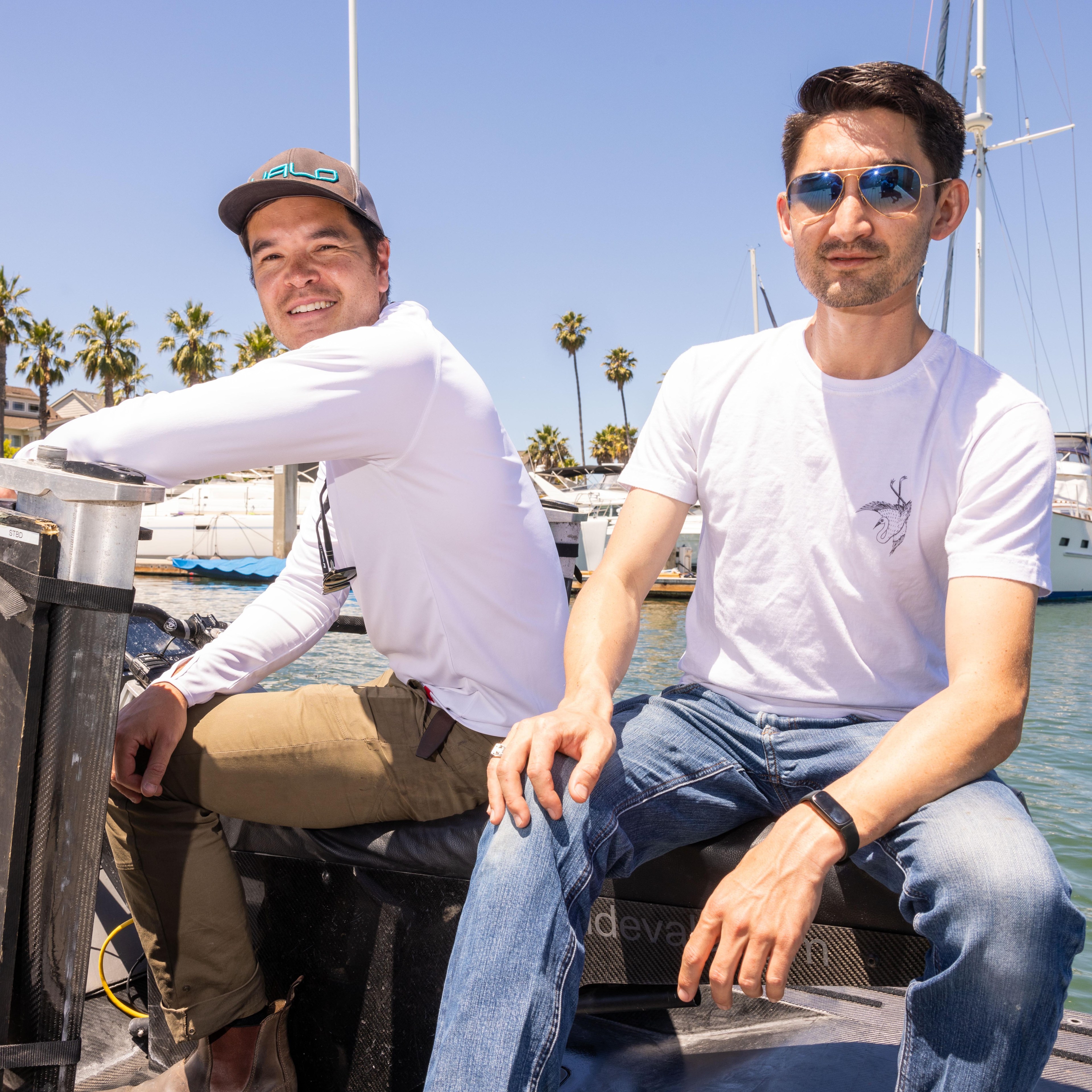 Two men are sitting on a boat docked at a marina, with yachts and palm trees in the background. One wears a cap and white long-sleeve, the other sunglasses and a white t-shirt.