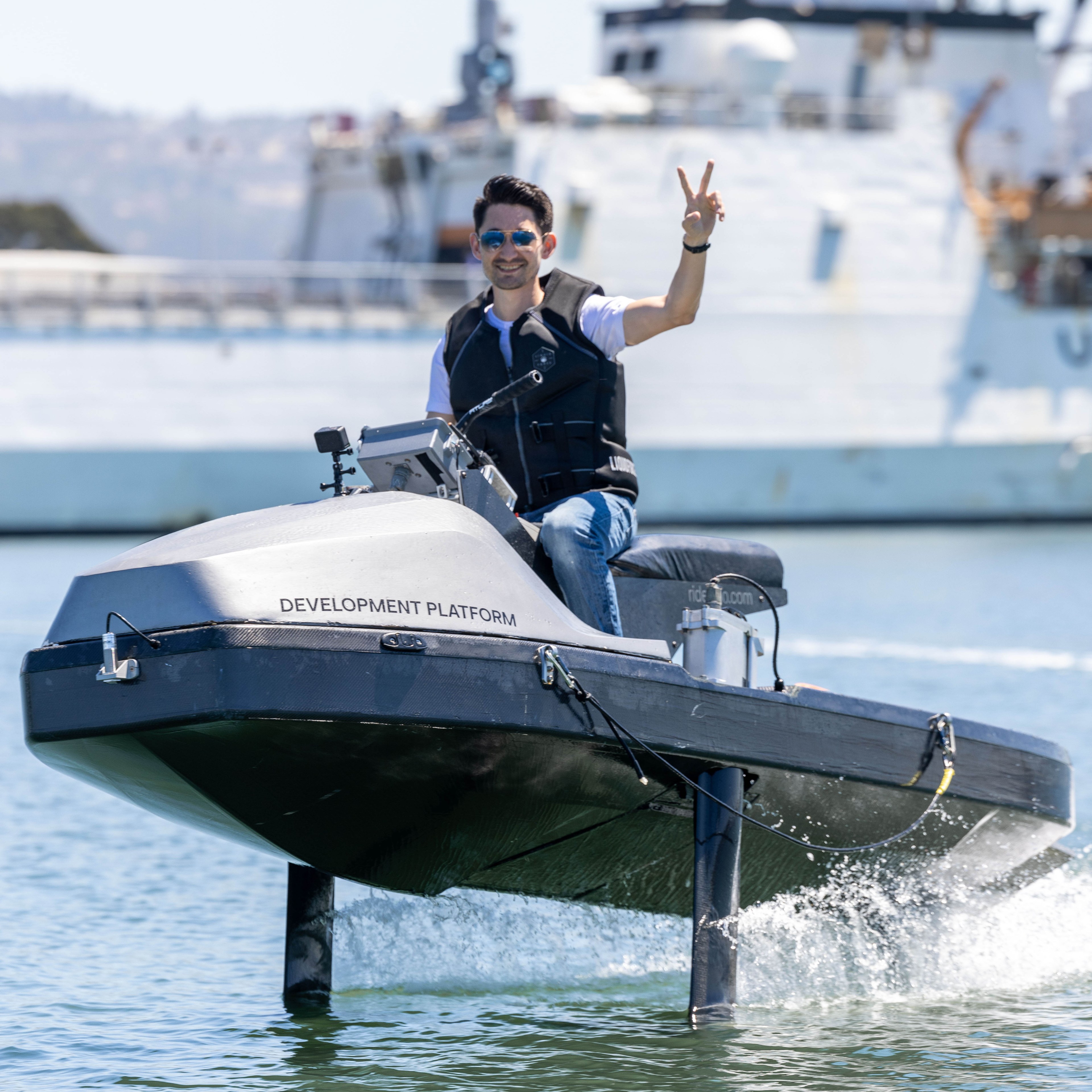 A man wearing sunglasses and a life vest smiles and makes a peace sign while riding a &quot;Development Platform&quot; hydrofoil boat on the water, with ships in the background.