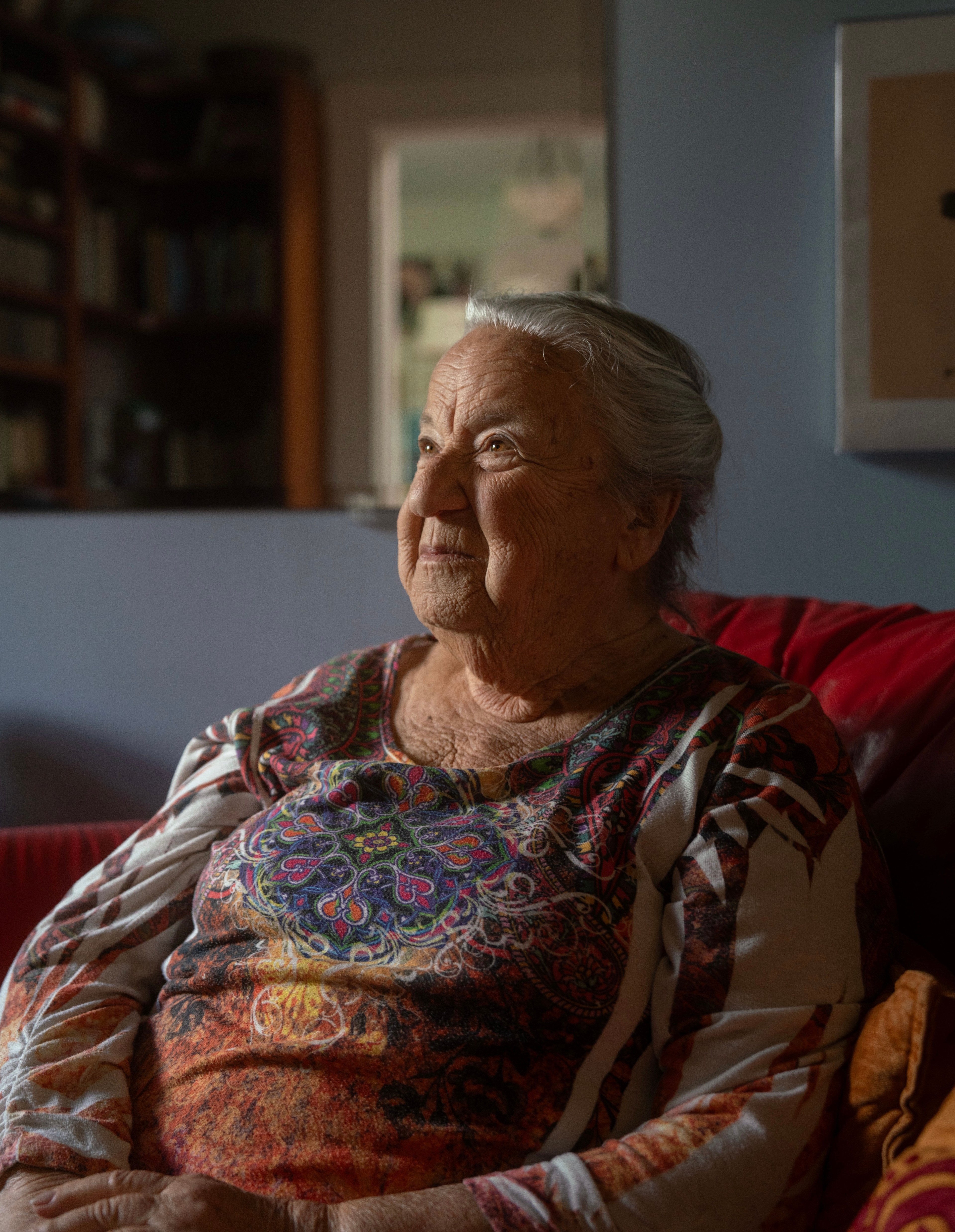 An elderly woman with gray hair sits in a cozy room, wearing a colorful, patterned shirt, looking thoughtfully to the side with a gentle expression.