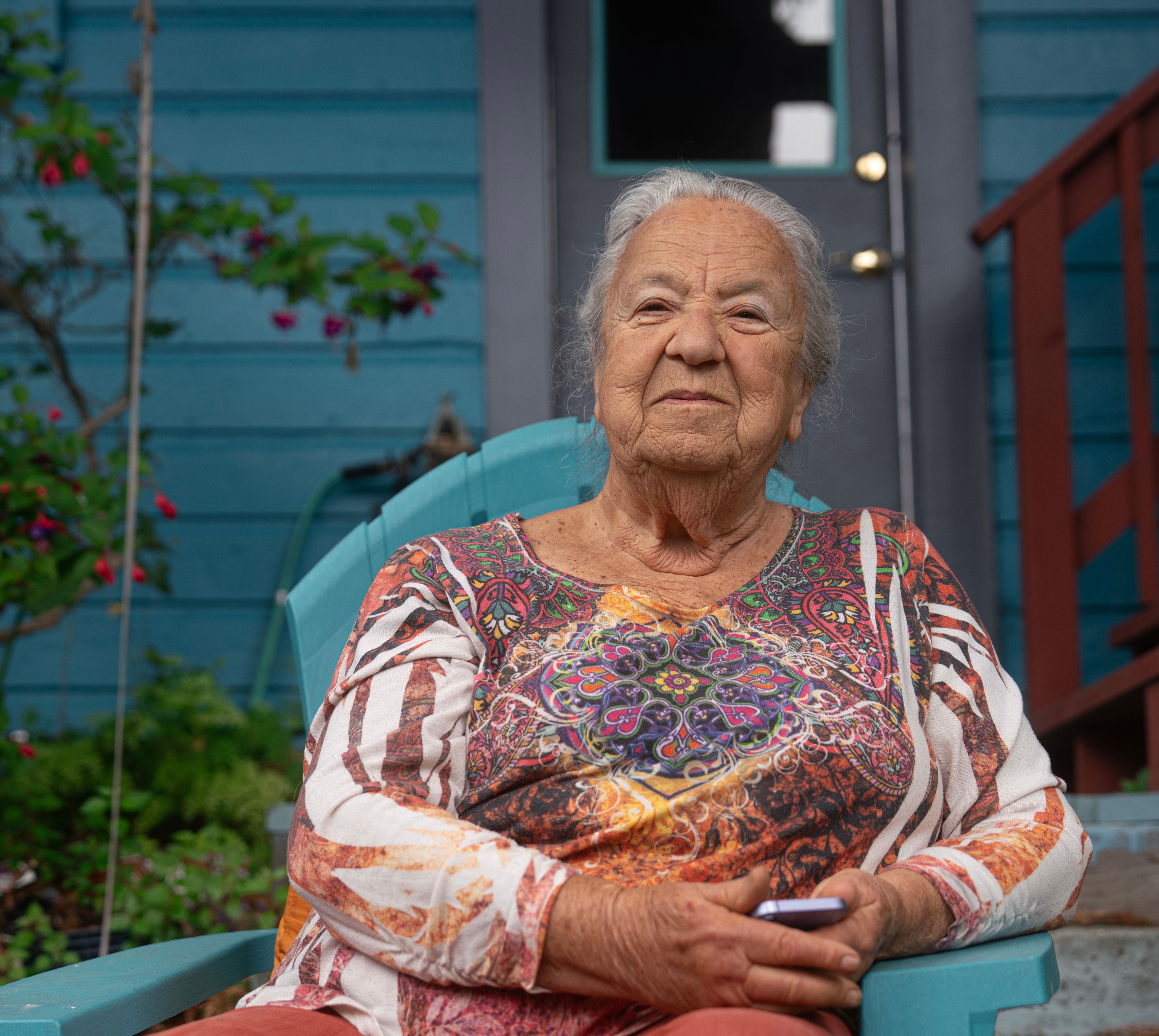 An elderly woman with white hair is sitting in a light blue chair. She is wearing a colorful, patterned top and holding a phone. Behind her is a blue house with a red railing.