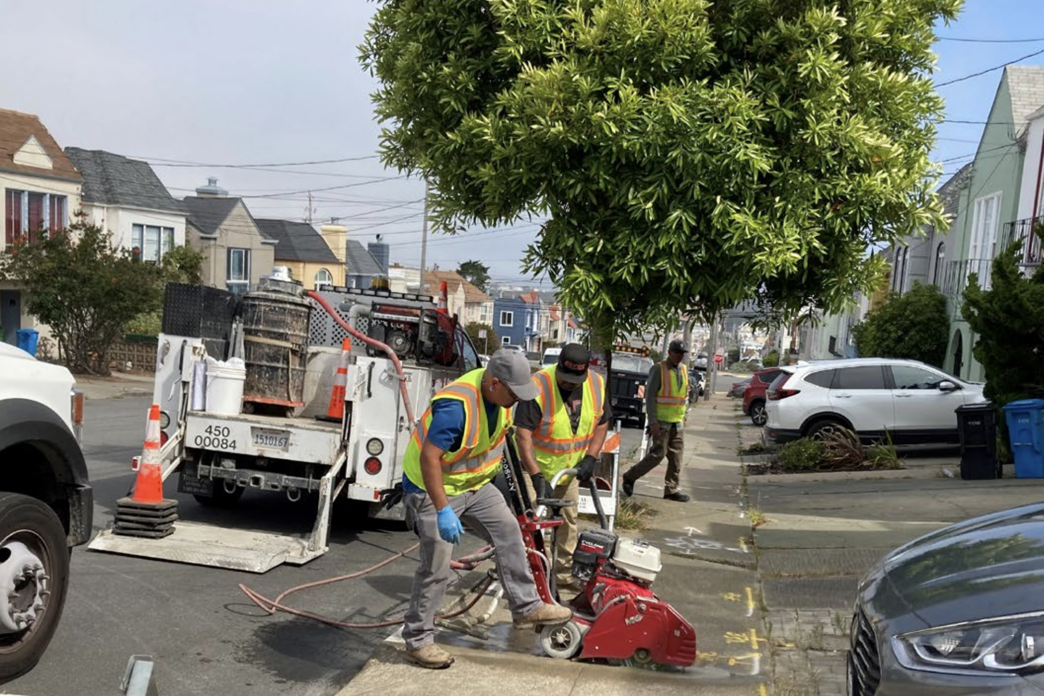 Construction workers in safety vests are operating machinery and working on a sidewalk in a residential neighborhood with parked cars and houses visible in the background.