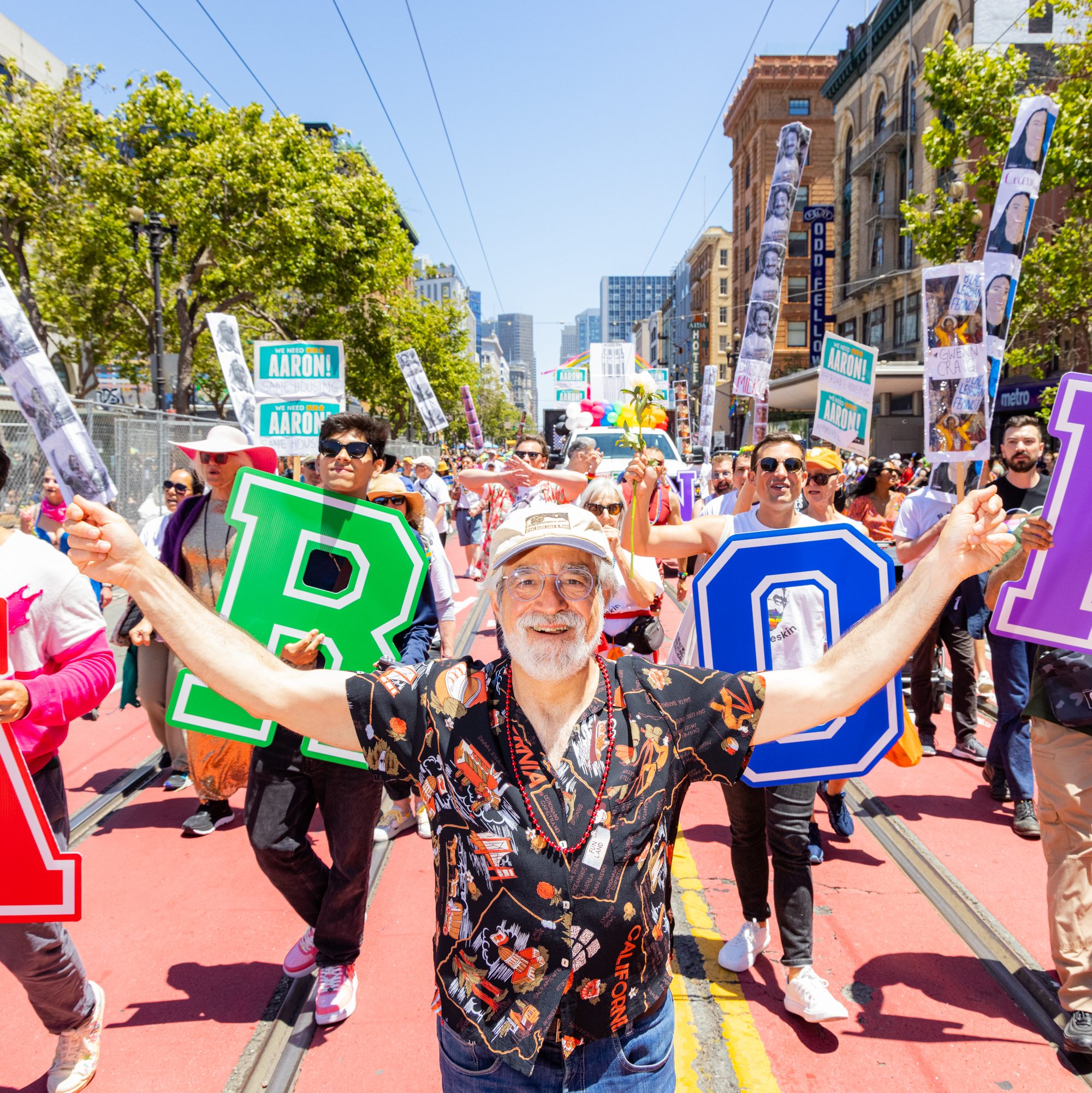 A joyous man leads a colorful street parade, holding large foam letters. Behind him, people wave signs and march along a sunny, vibrant city street.