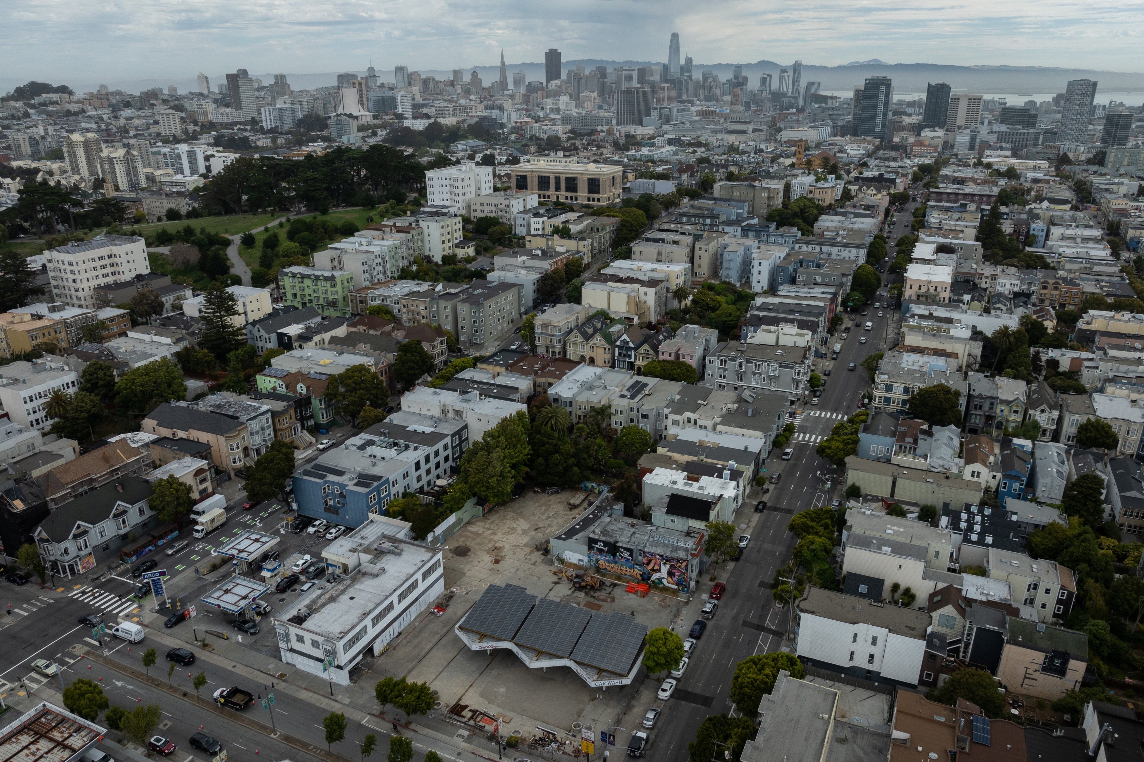 The image shows an aerial view of a dense urban neighborhood with a mix of low-rise buildings, greenery, and a distant city skyline under a cloudy sky.