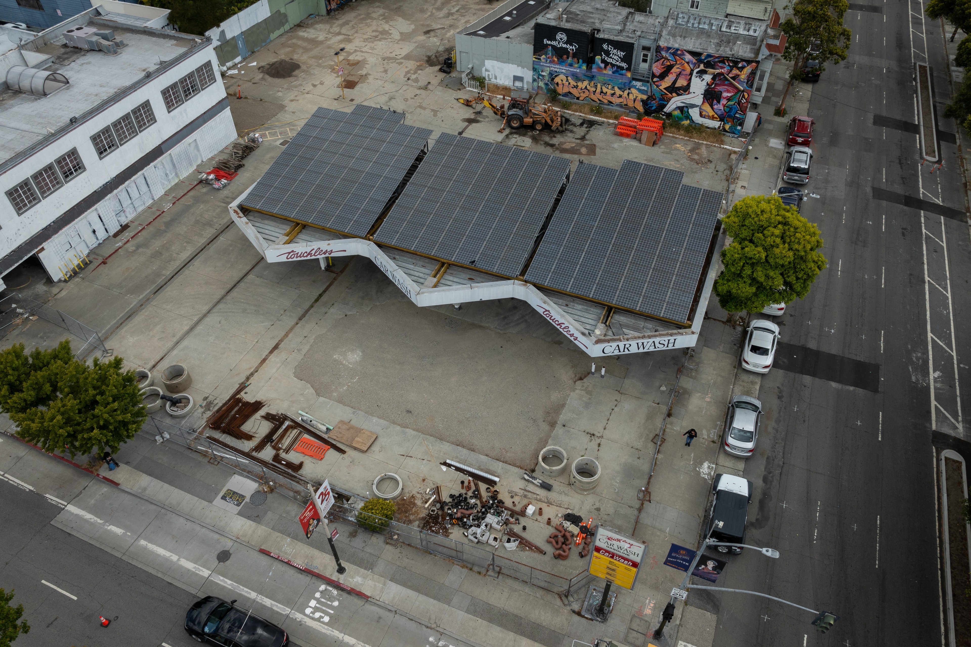An aerial view shows a car wash with solar panels on its roof, surrounded by an industrial area, a road with parked cars, and some graffiti art on adjacent buildings.