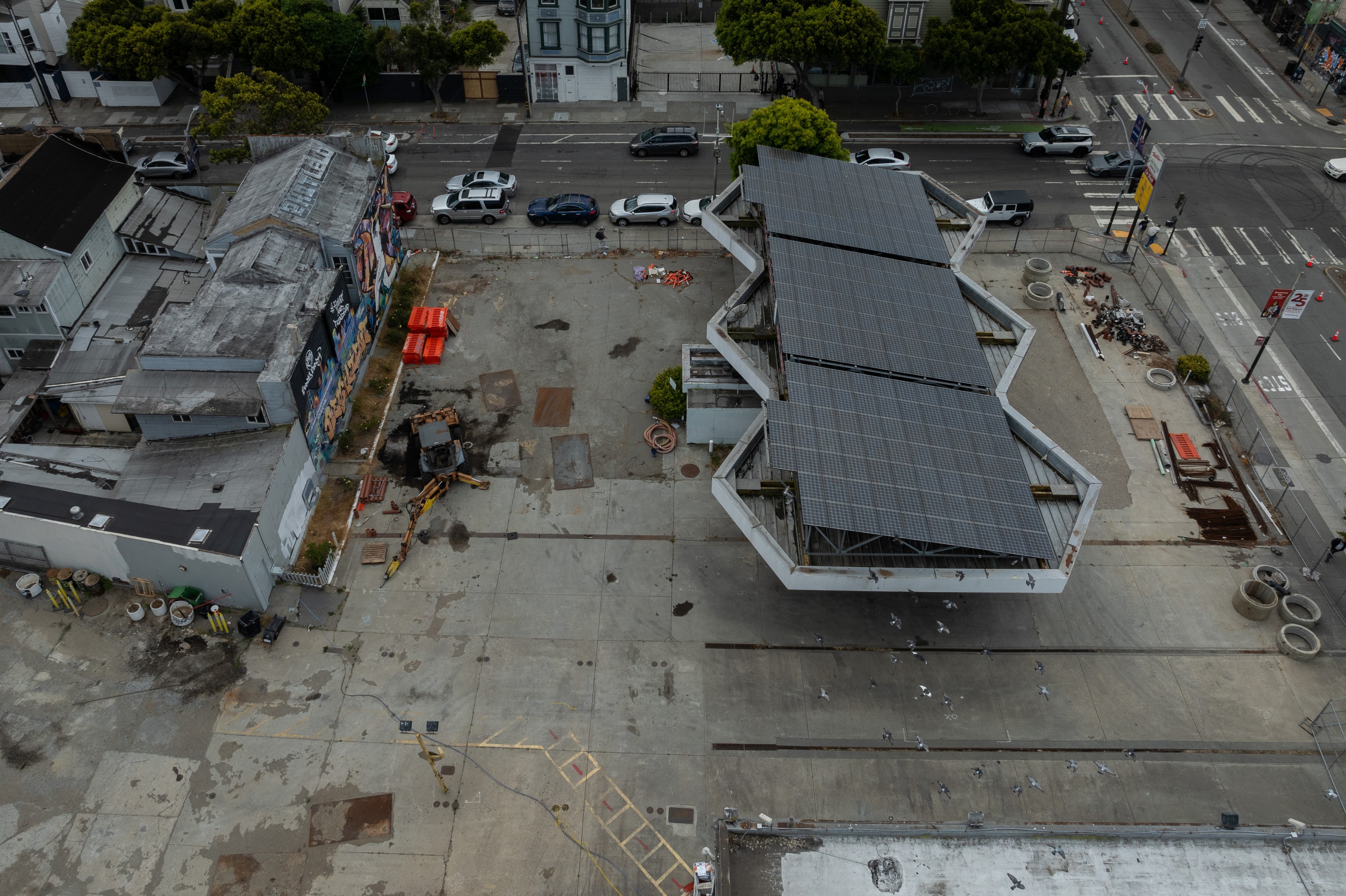 An aerial view shows an urban area with parked cars along a road, an old building with a colorful mural, a partially constructed space with scattered materials, and solar panels.