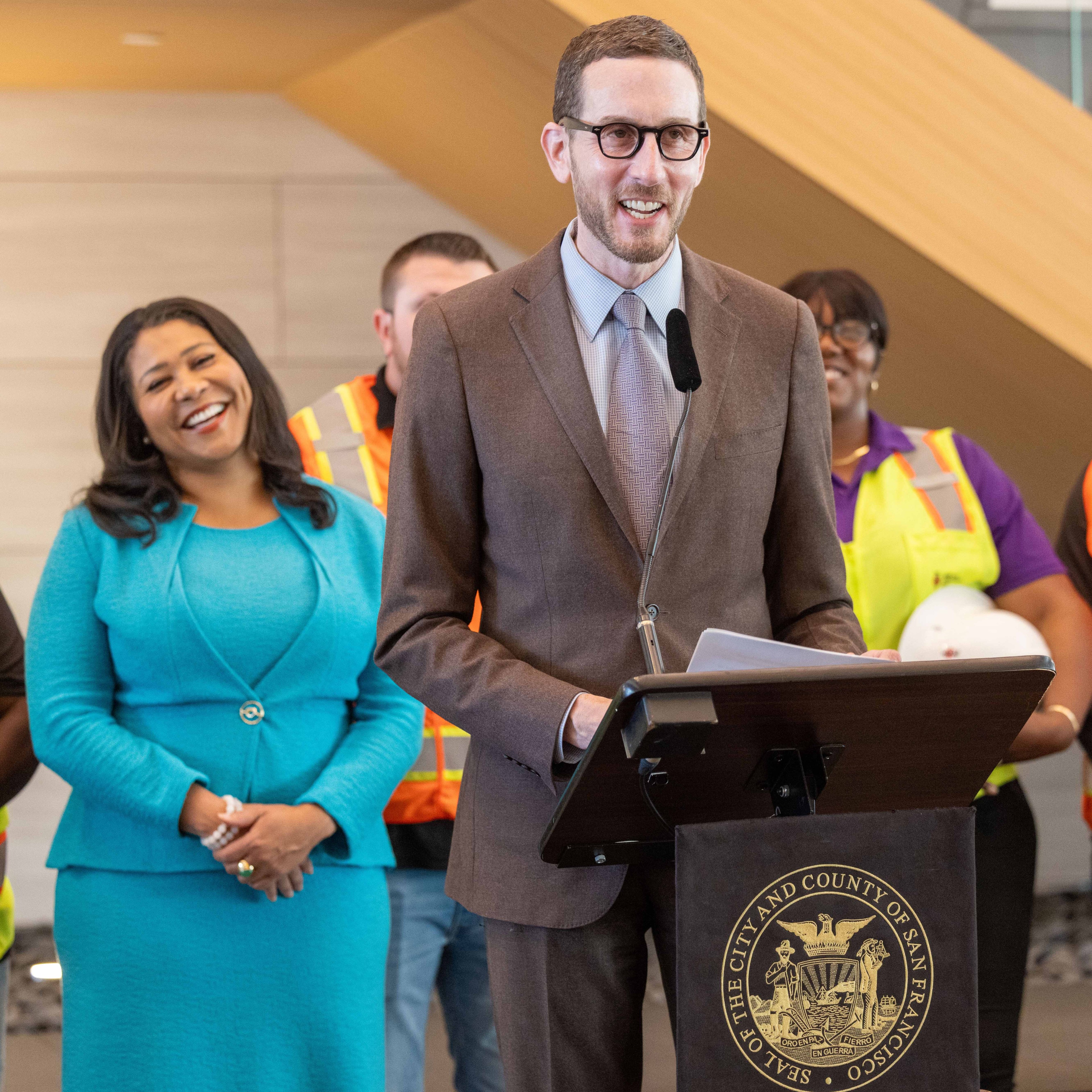 A man in a brown suit speaks at a podium with a microphone, while a woman in a blue outfit smiles behind him. Several people in construction attire stand in the background.