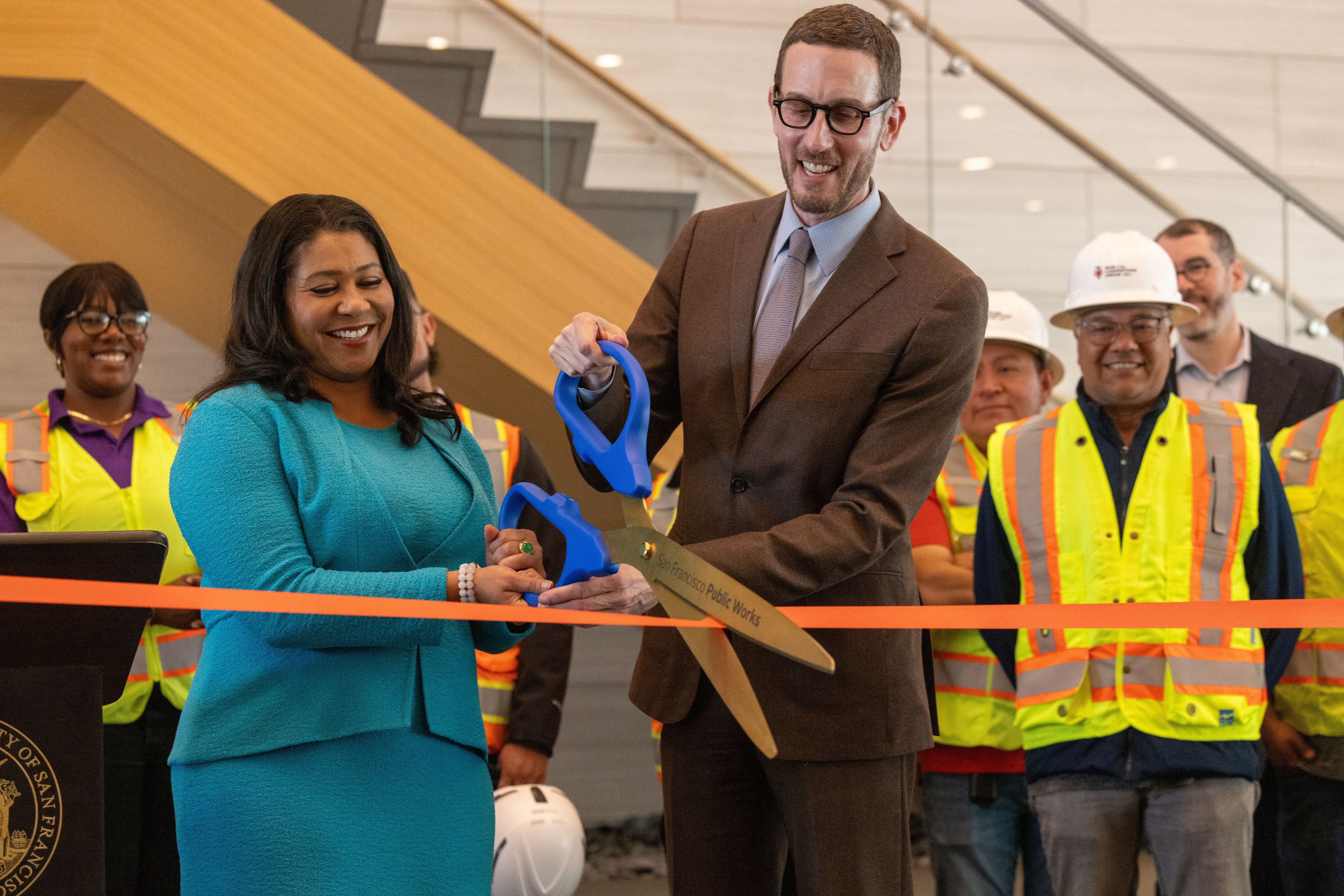 Two people in formal attire are smiling while cutting a red ribbon with oversized blue scissors, surrounded by construction workers wearing yellow vests and helmets.