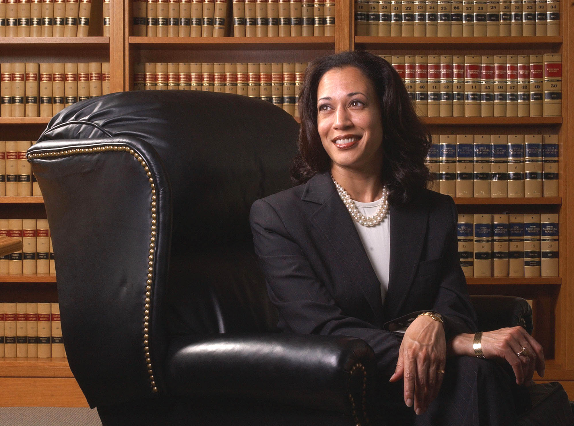 A woman in a dark suit and pearl necklace sits in a leather chair, smiling. Behind her, shelves filled with legal books are visible, creating a scholarly setting.