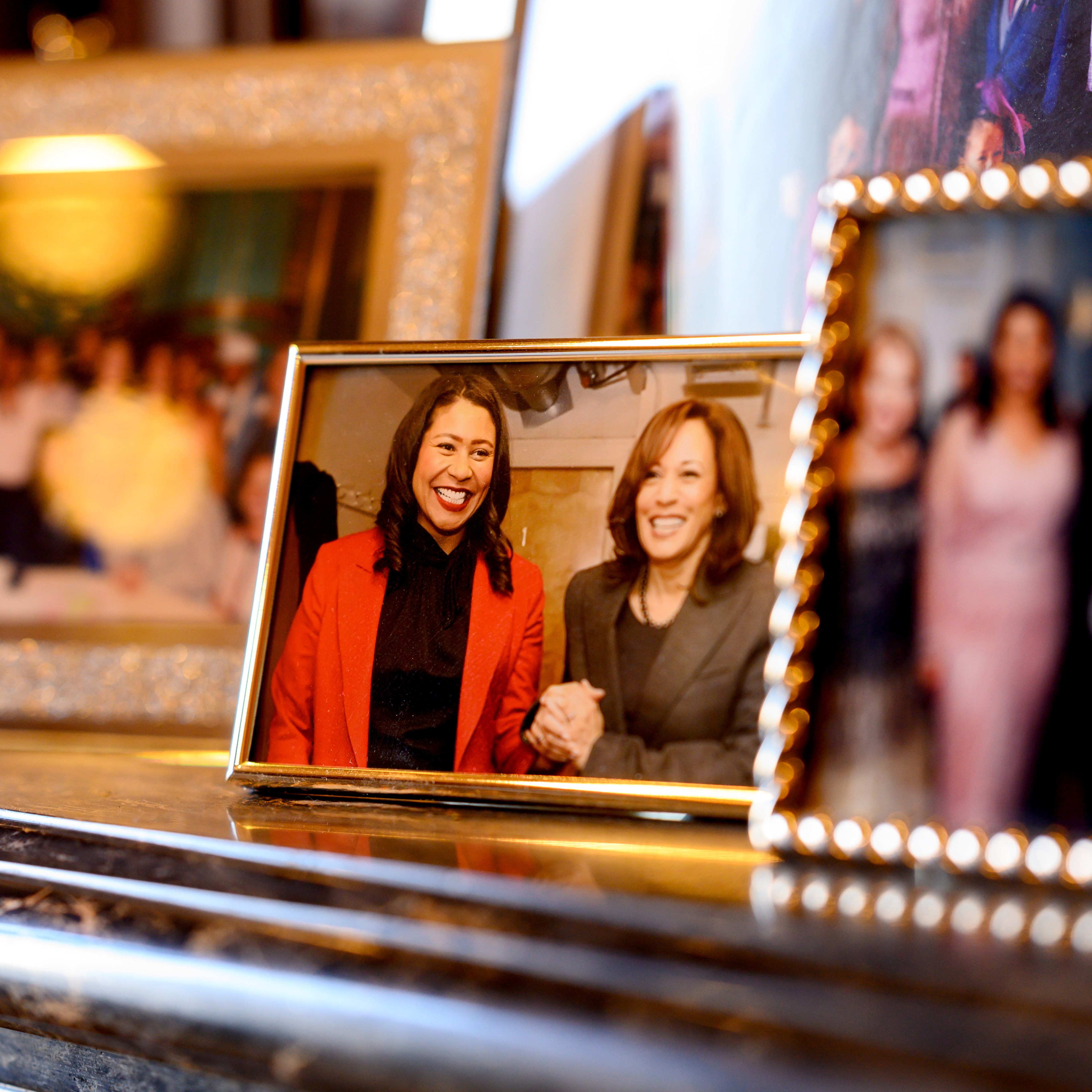 The image shows several framed photographs with the central one featuring two smiling women holding hands, dressed in professional attire. The setting appears to be indoors.