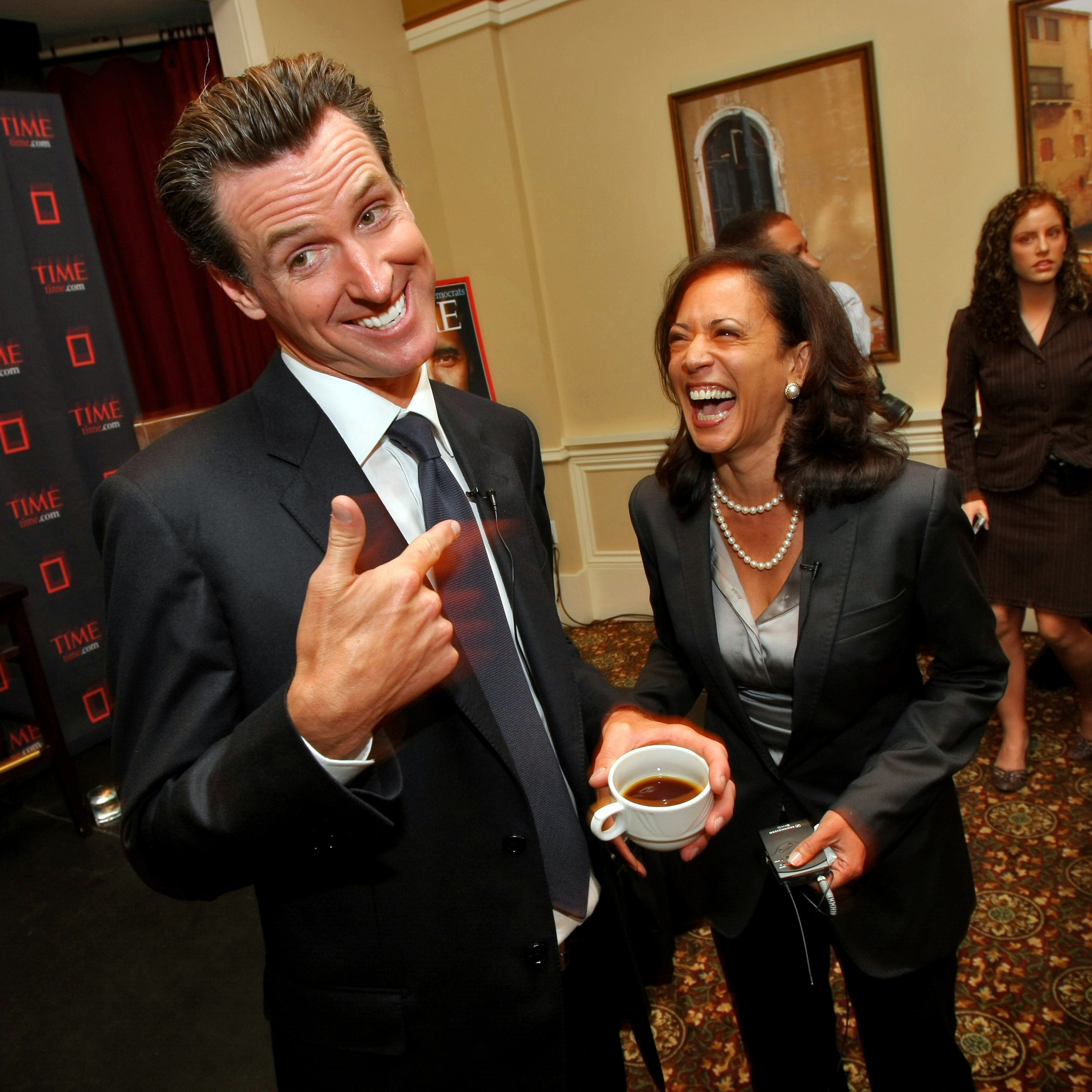 Two people in suits are laughing; one points to the other while holding a coffee cup. A &quot;TIME&quot; banner is in the background and another person is in view.