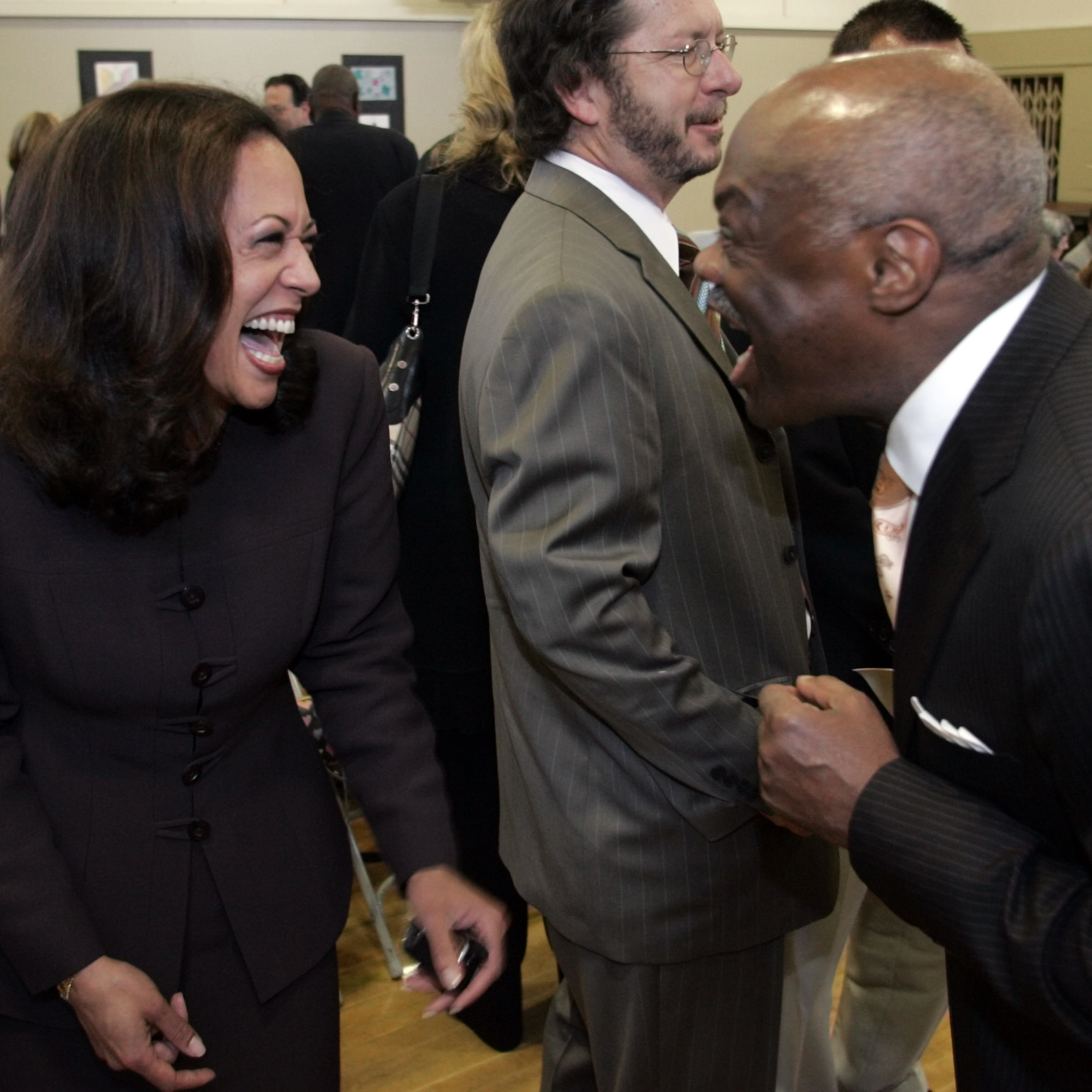 Two people are laughing and enjoying a lively conversation, both dressed in business attire. A man with glasses stands between them, appearing neutral.