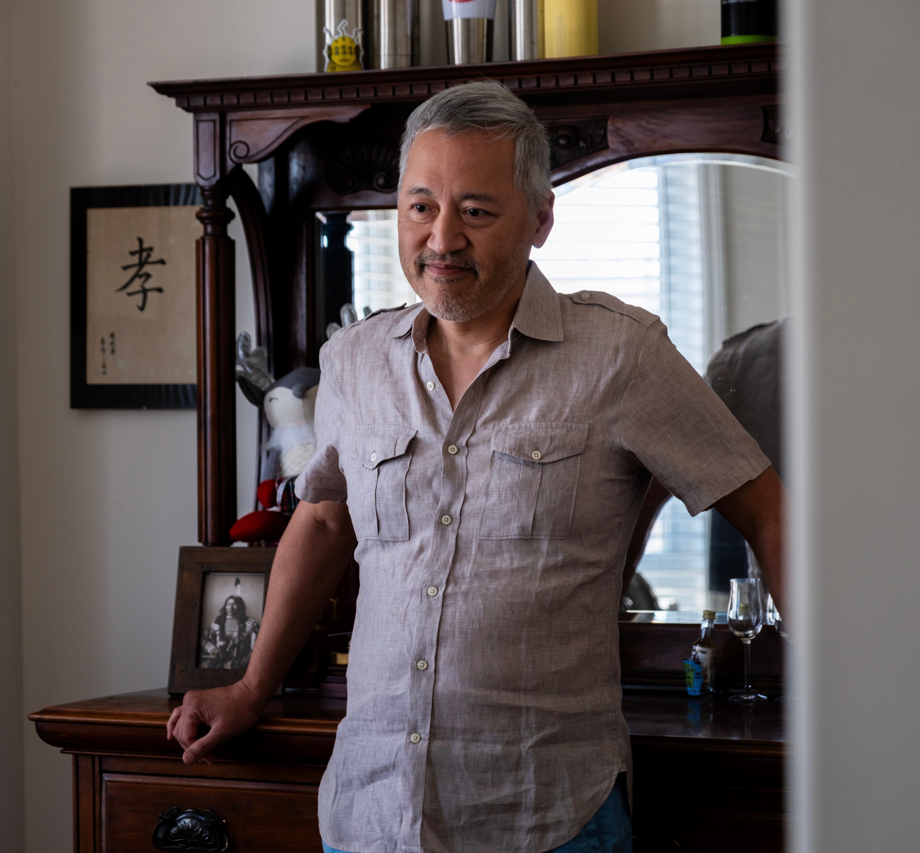 A man with gray hair, wearing a beige short-sleeved shirt, stands near a wooden dresser with a mirror. Behind him are framed art, a plush toy, and various items on the dresser.