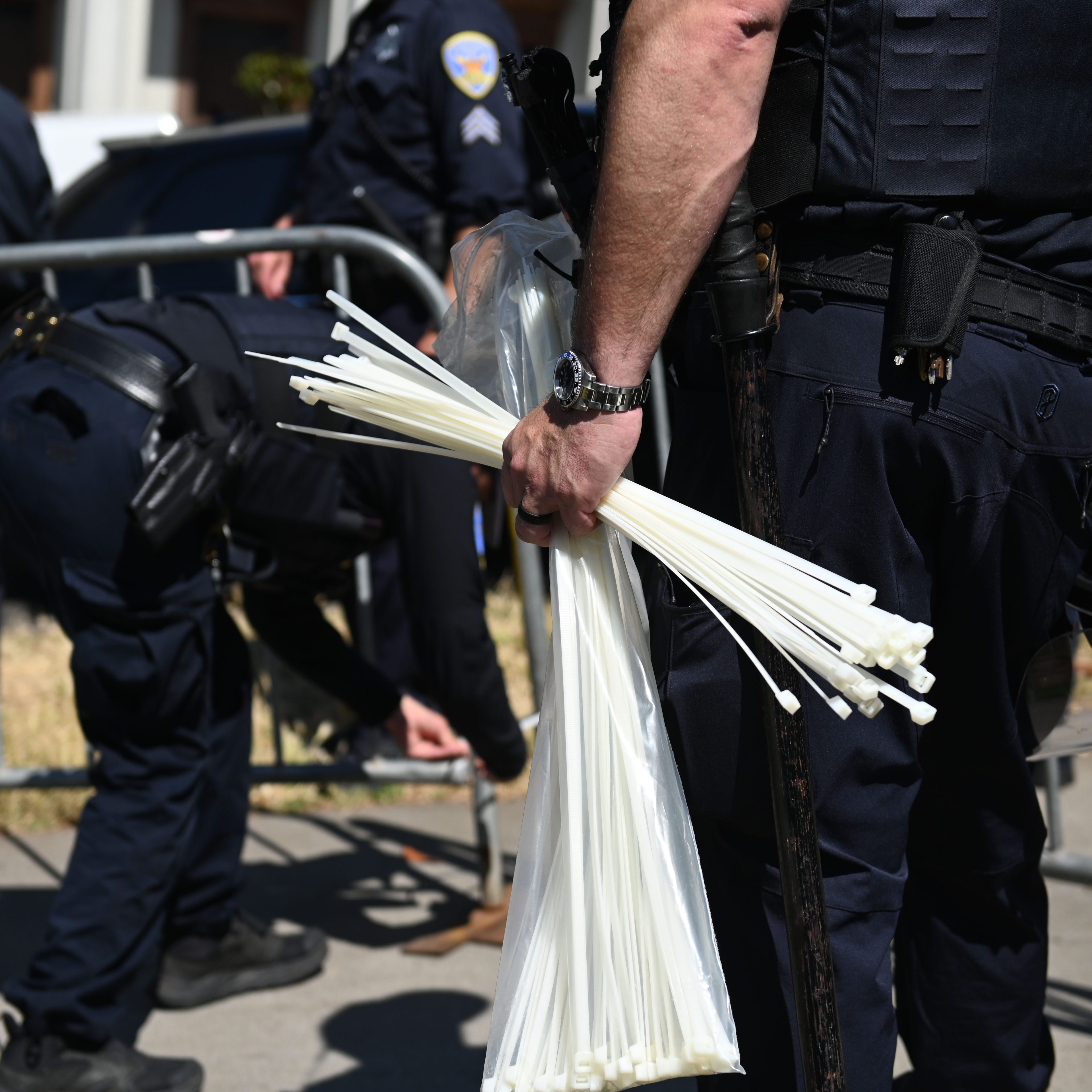 Police officers in dark uniforms are standing near a barricade. One officer is holding a bundle of large white zip ties, while another officer bends down by the barricade.