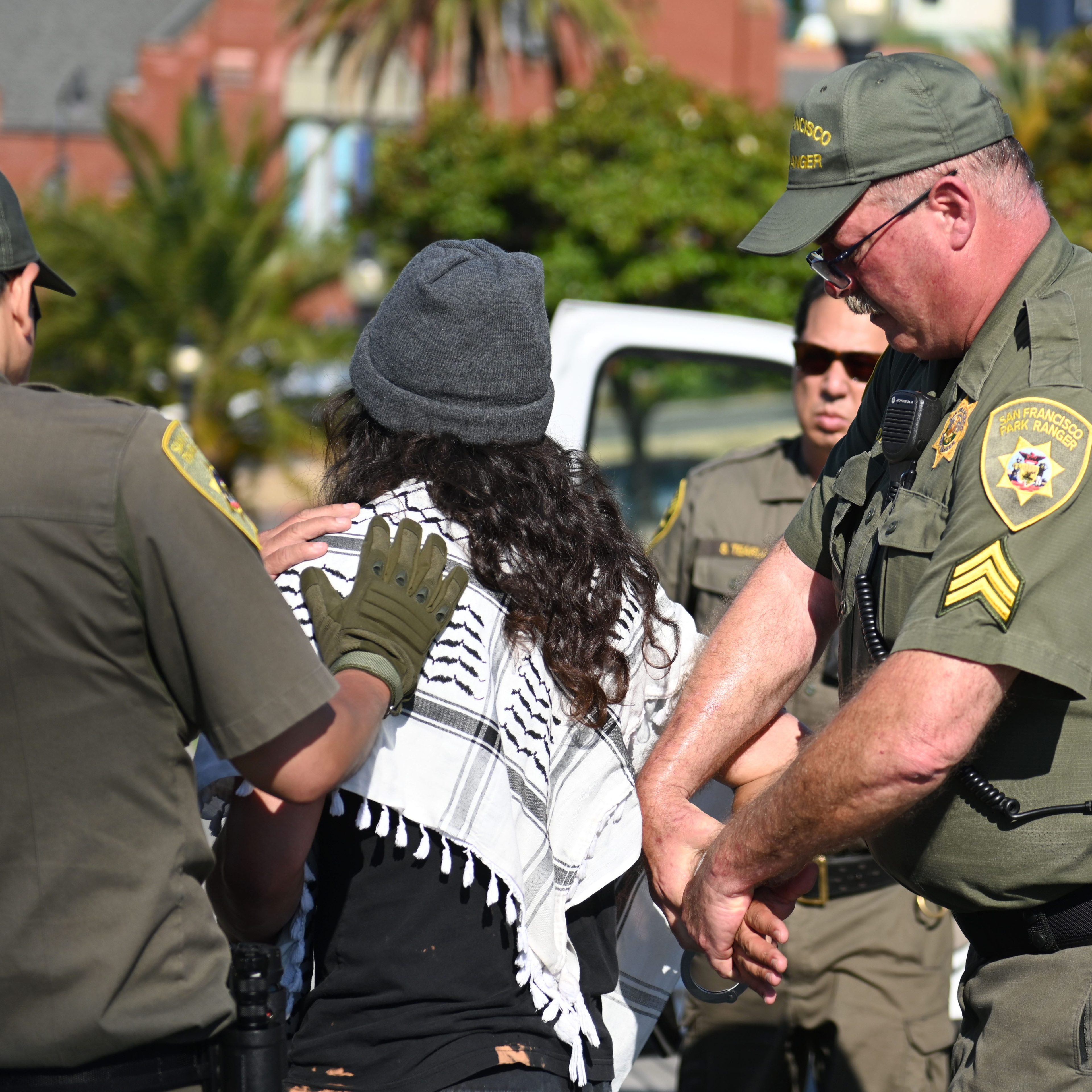 Three park rangers are arresting a person in a beanie and patterned scarf in front of a parked vehicle with a cage on the back.