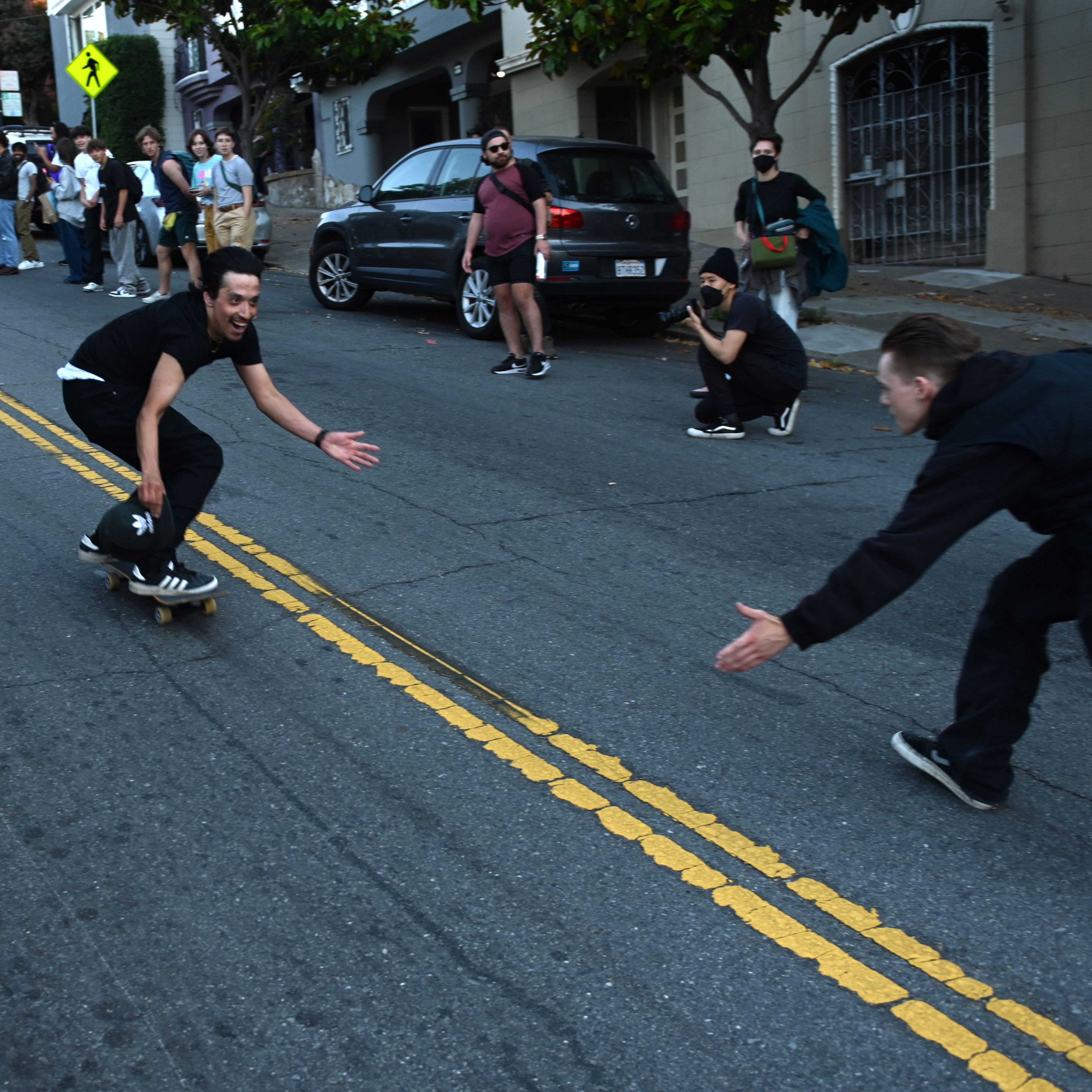 A skateboarder rides down a street, reaching out to another person extending their hand. Onlookers and photographers line the sidewalks, capturing the action.