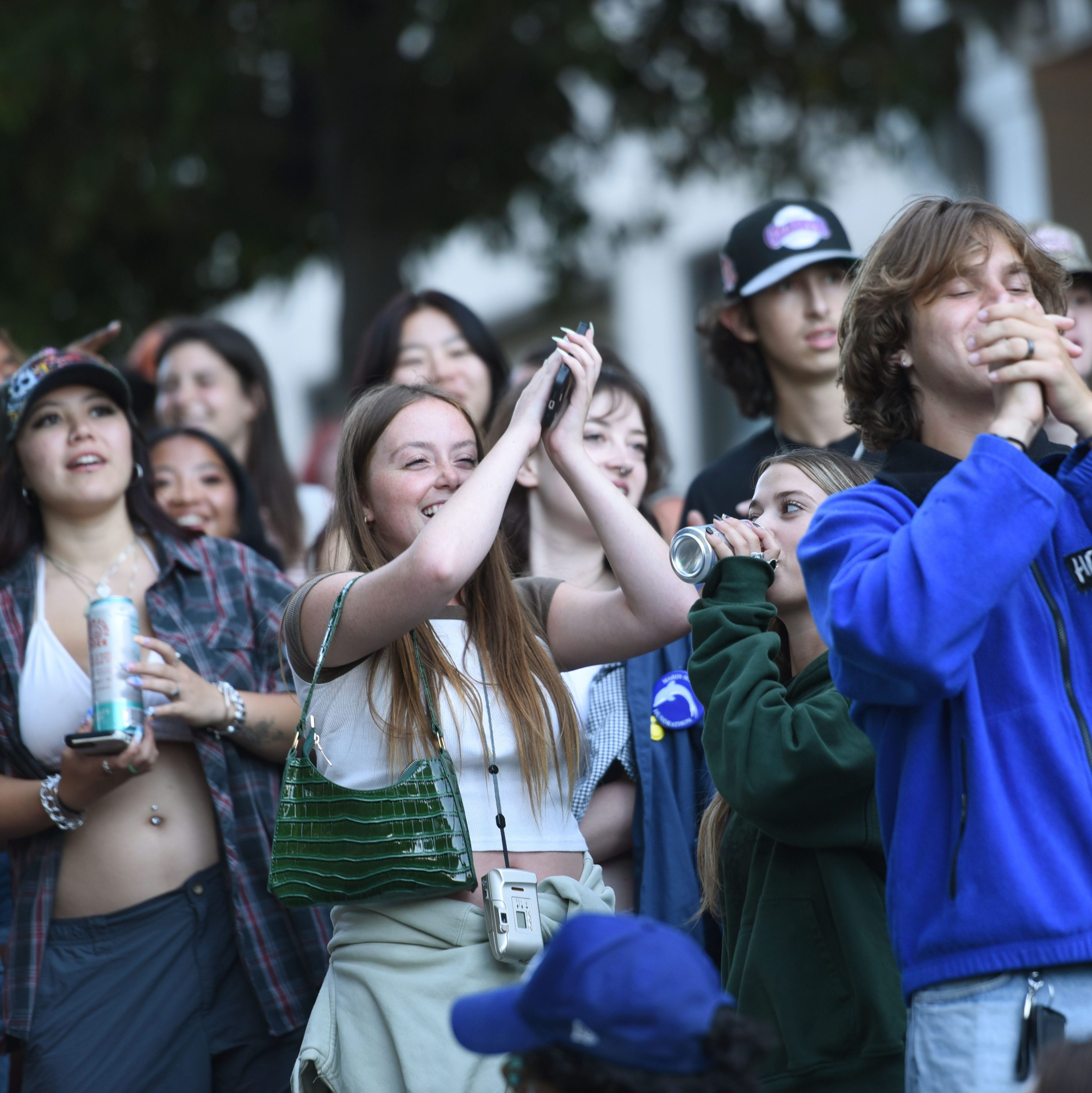 A group of young people, mostly in casual wear, is gathered outdoors. They appear excited, some clapping, others cheering, with a few holding cans and phones. Trees are in the background.