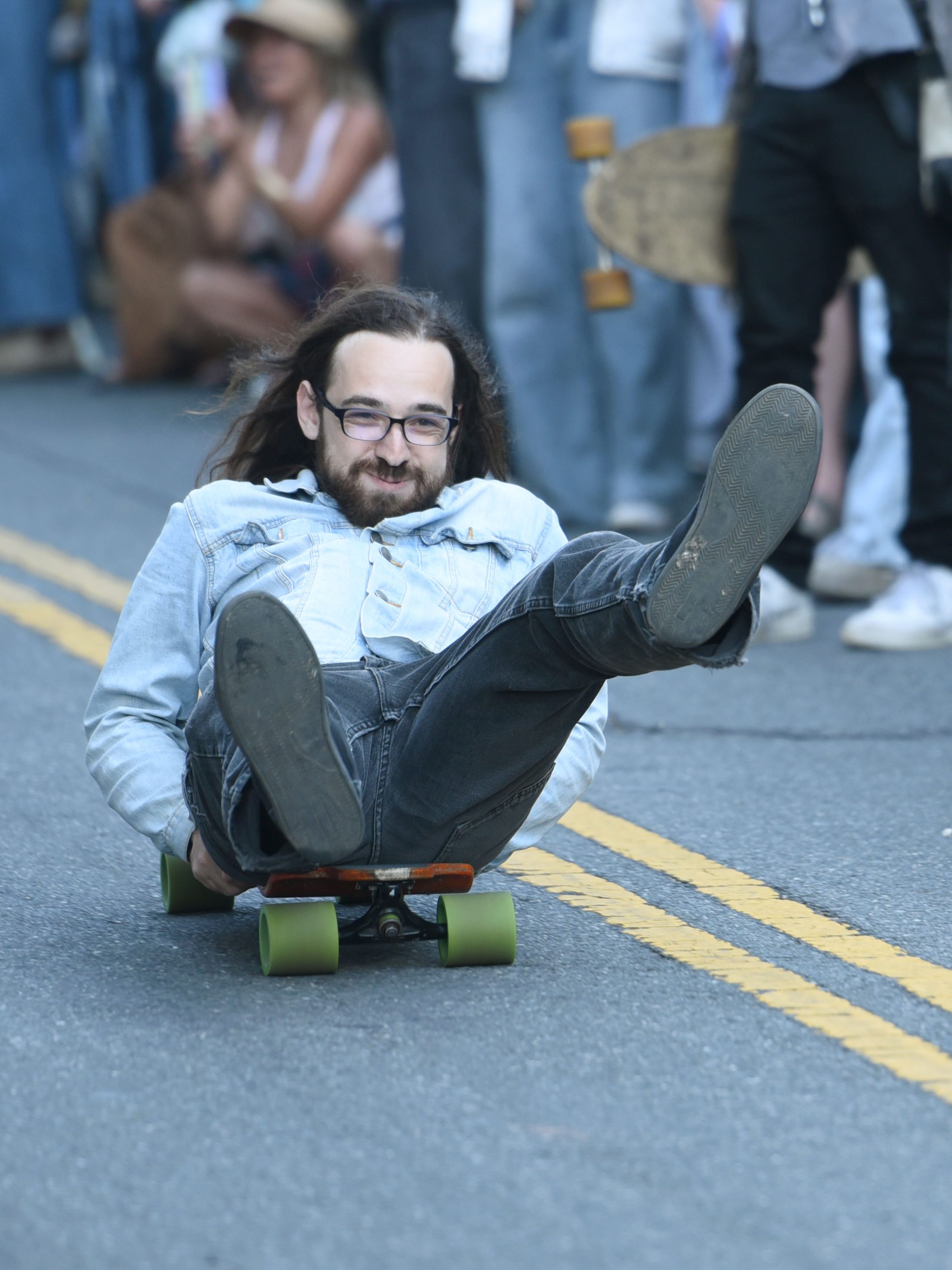 A man with long hair is lying on a skateboard, riding down the middle of a street surrounded by standing spectators. He appears to be enjoying the ride.