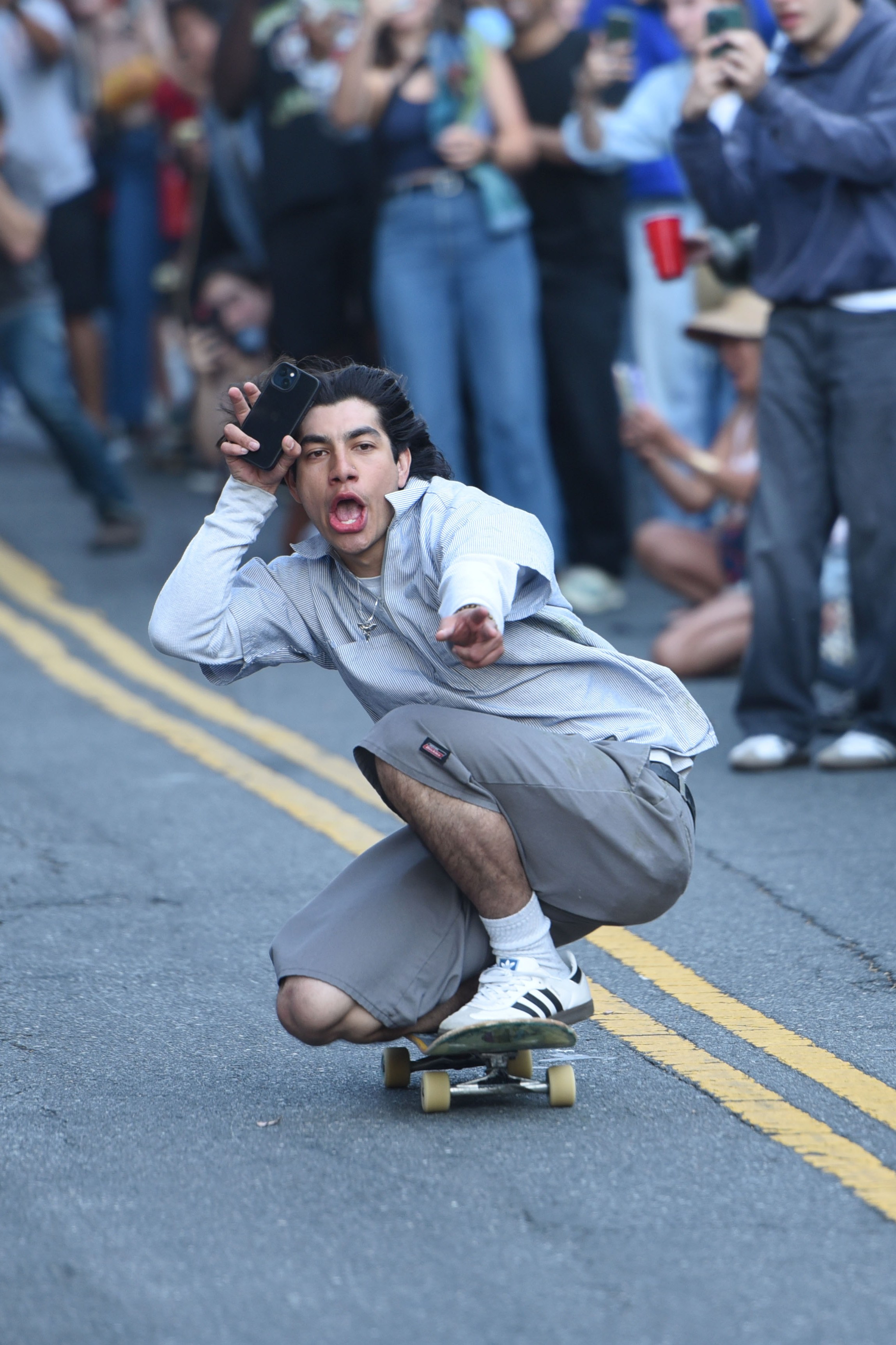 A man skateboards down a street, crouching low with a playful expression, while holding a phone up. A crowd of onlookers, many taking photos, lines both sides of the street.