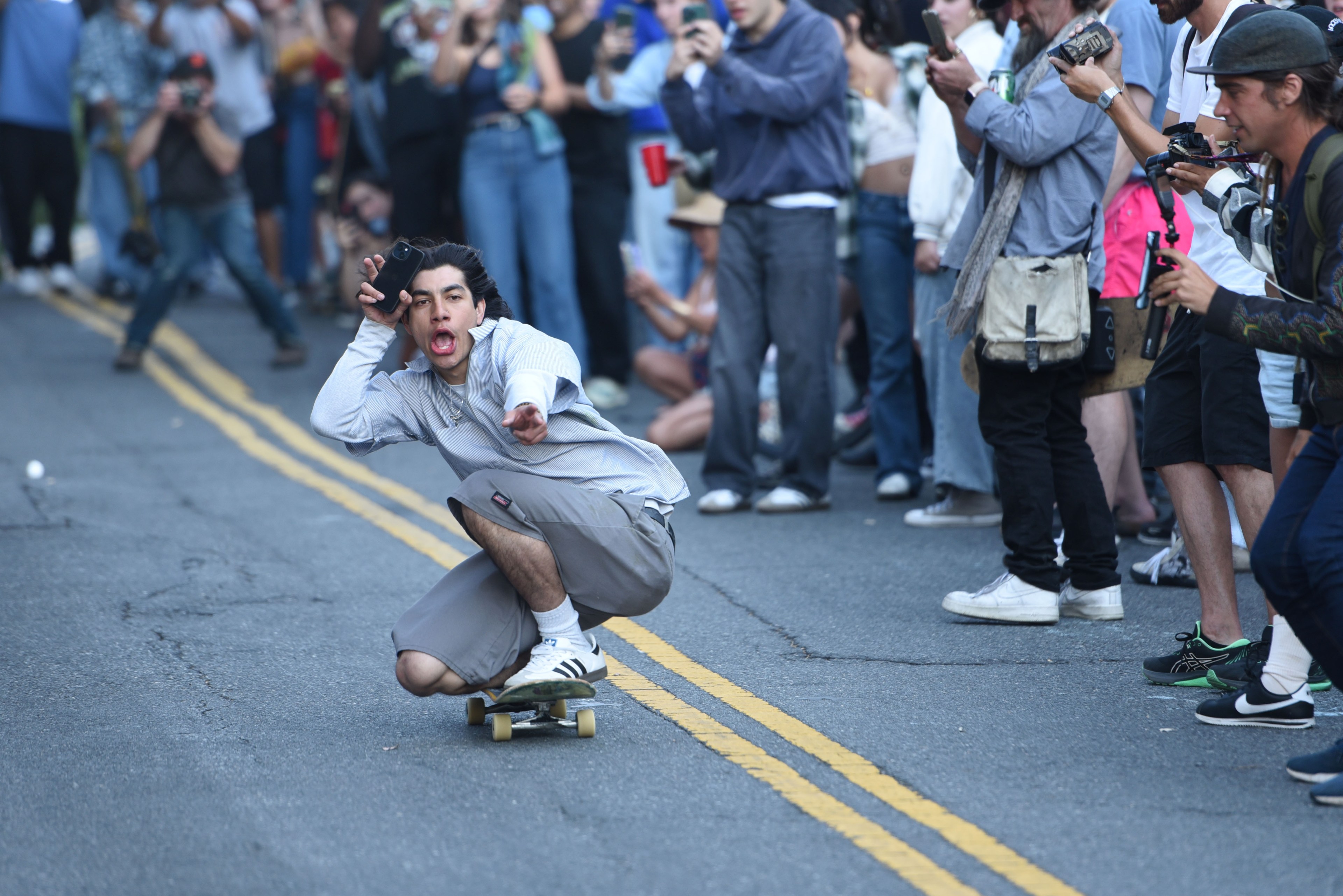 A man skateboards down a street, crouching low with a playful expression, while holding a phone up. A crowd of onlookers, many taking photos, lines both sides of the street.