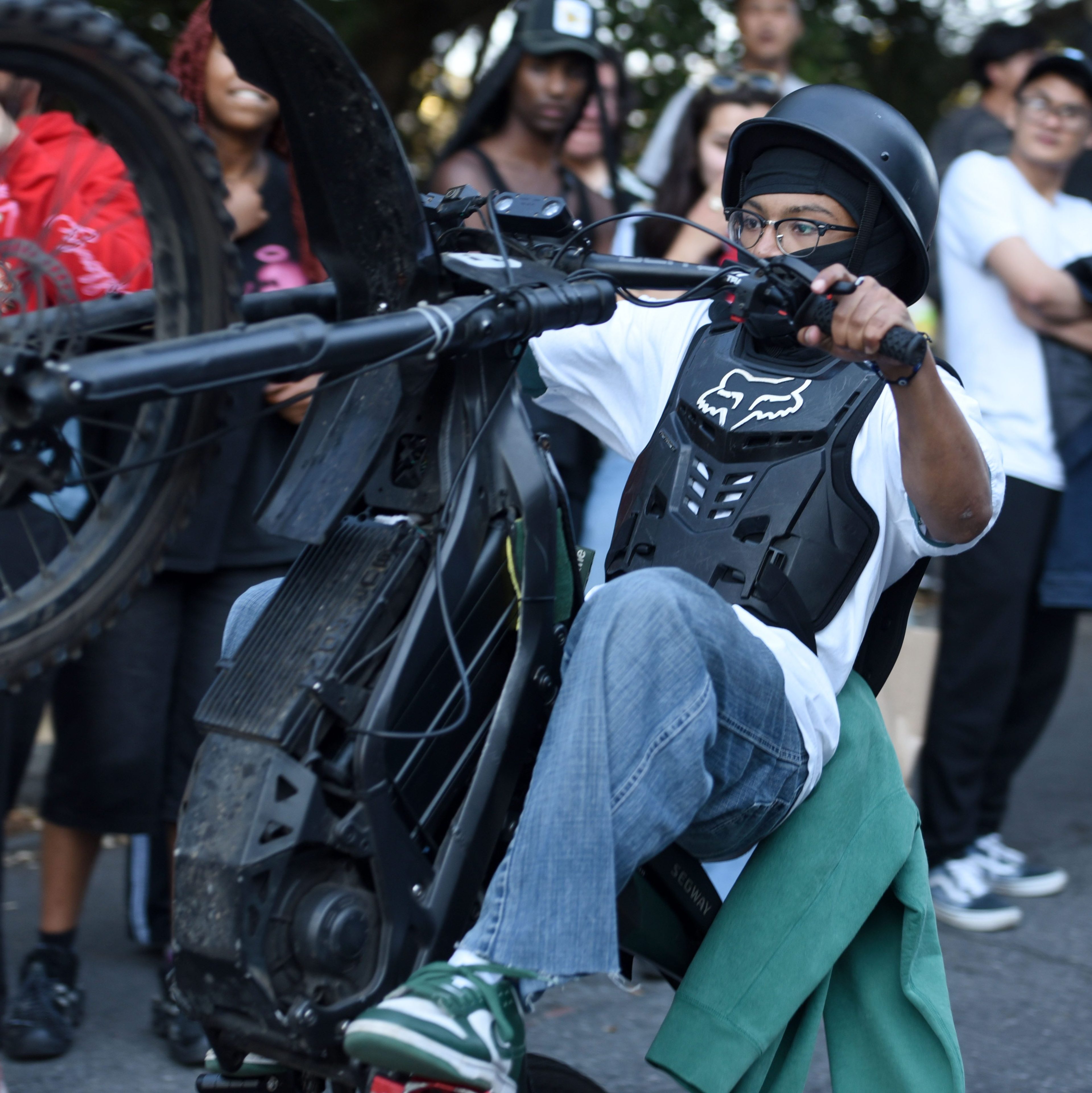 A person performs a wheelie on a mountain bike, wearing a helmet and protective gear, while a group of onlookers, some with cameras, watches and photographs.