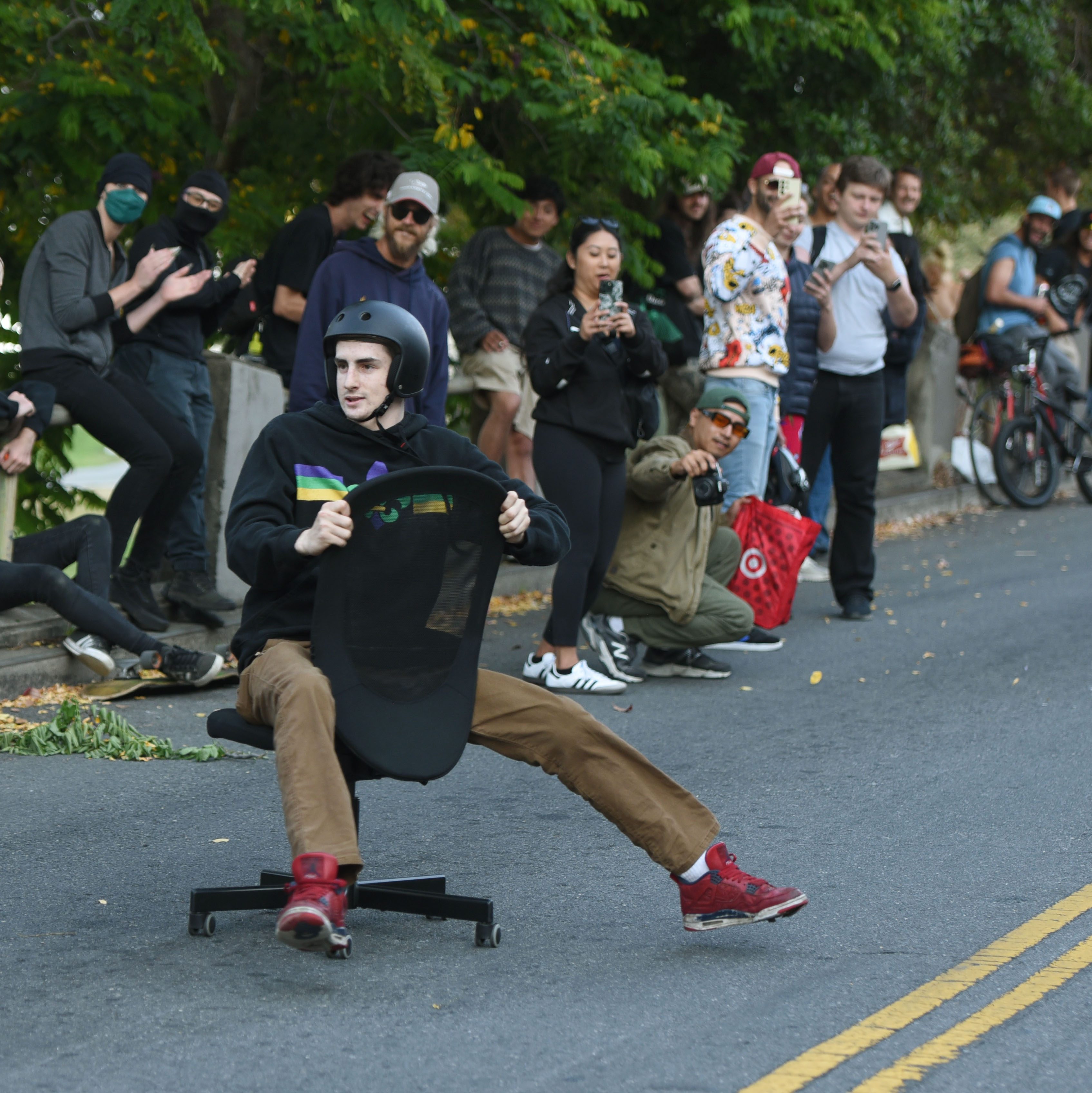 A man in a helmet rides an office chair down a street while a crowd of onlookers, some taking photos and videos, cheer him on from the roadside.