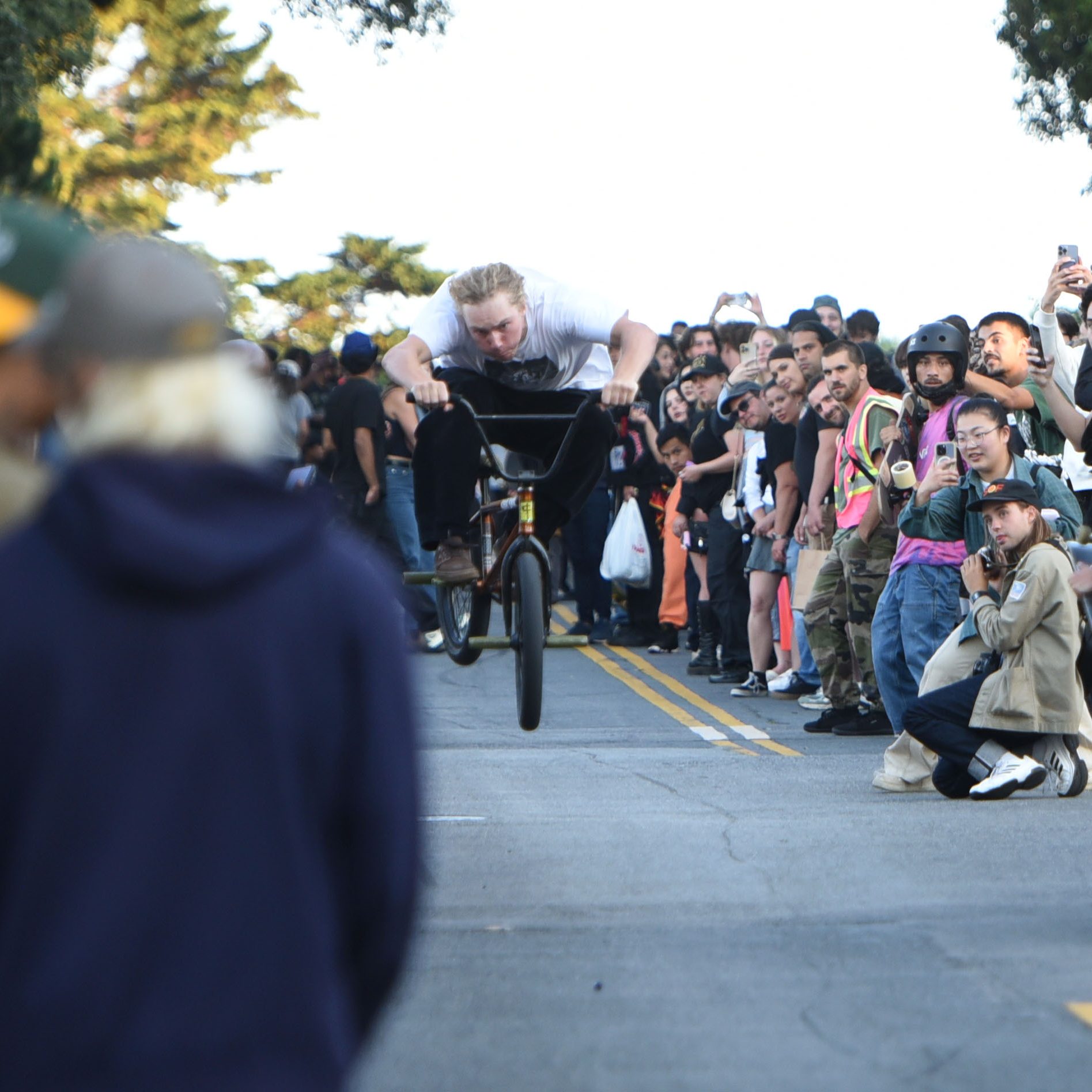 A cyclist, airborne on a bicycle, is performing a stunt on a street lined with spectators capturing the moment with their phones. The background features trees and clear skies.