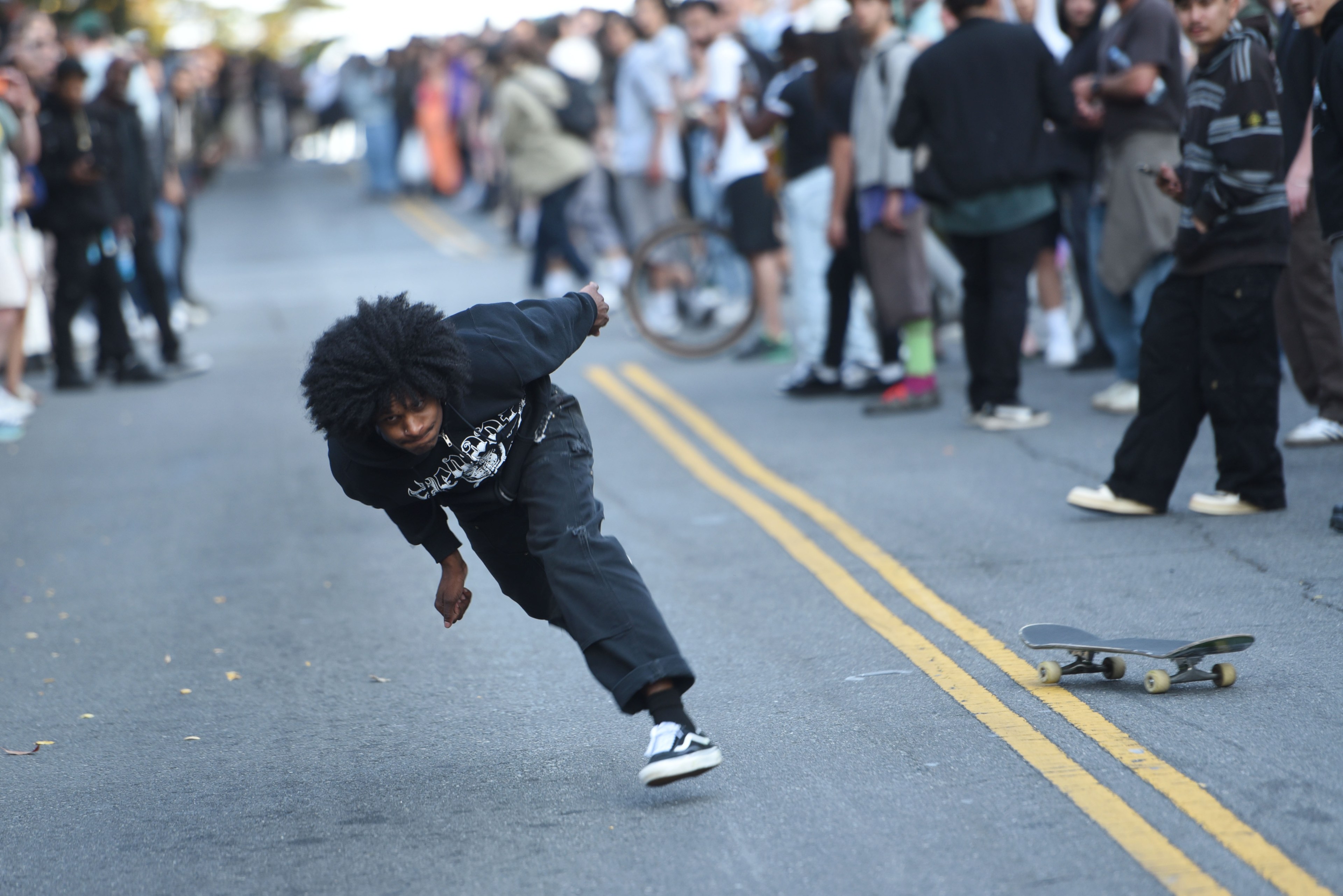 A person in a black hoodie and pants is falling while skateboarding on a street, with a skateboard on the ground. A crowd of people gathers in the background.