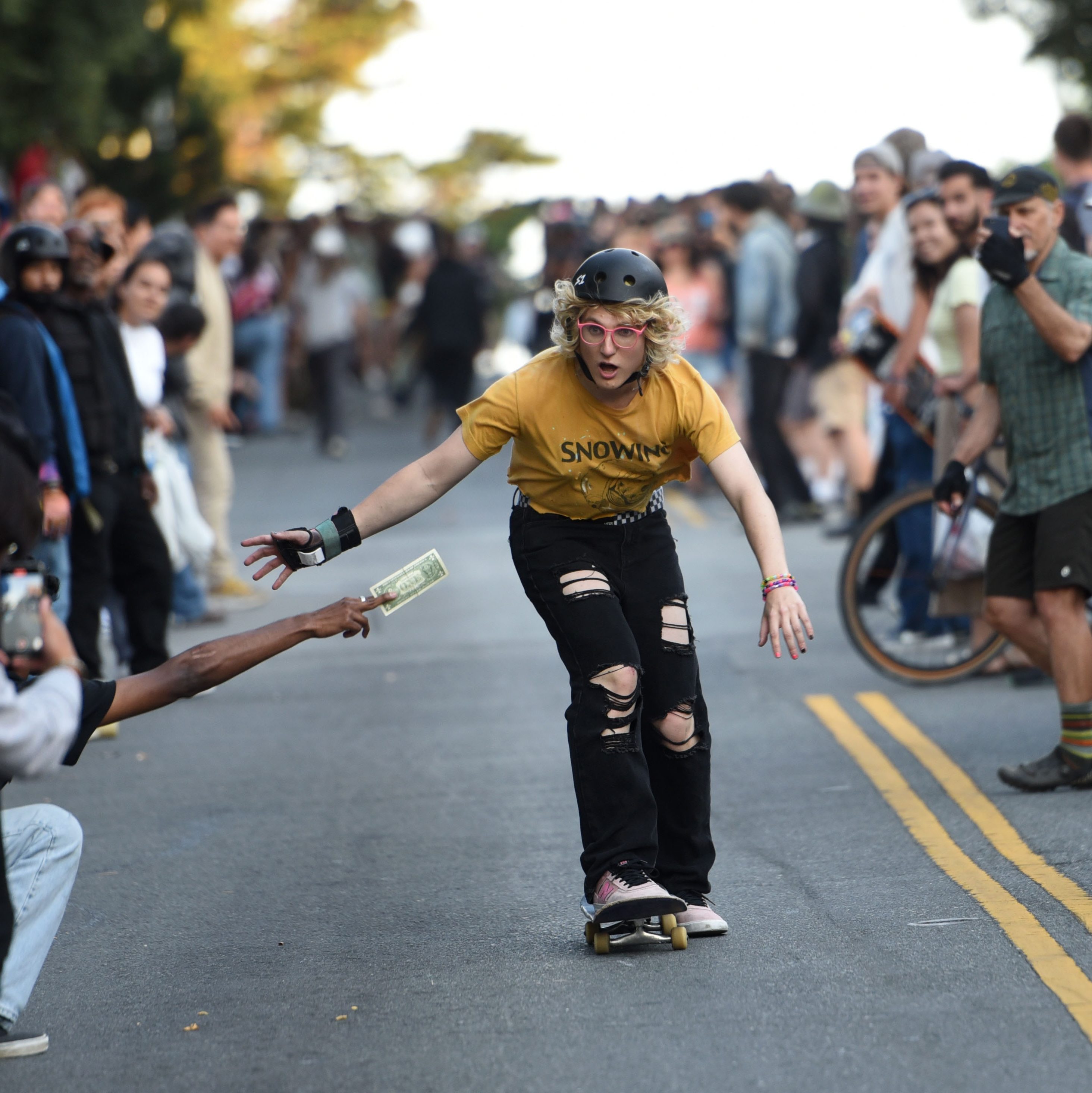 A skateboarder wearing a helmet and yellow shirt skates down a street, reaching for a dollar bill held out by a spectator. A crowd watches and a photographer kneels nearby.