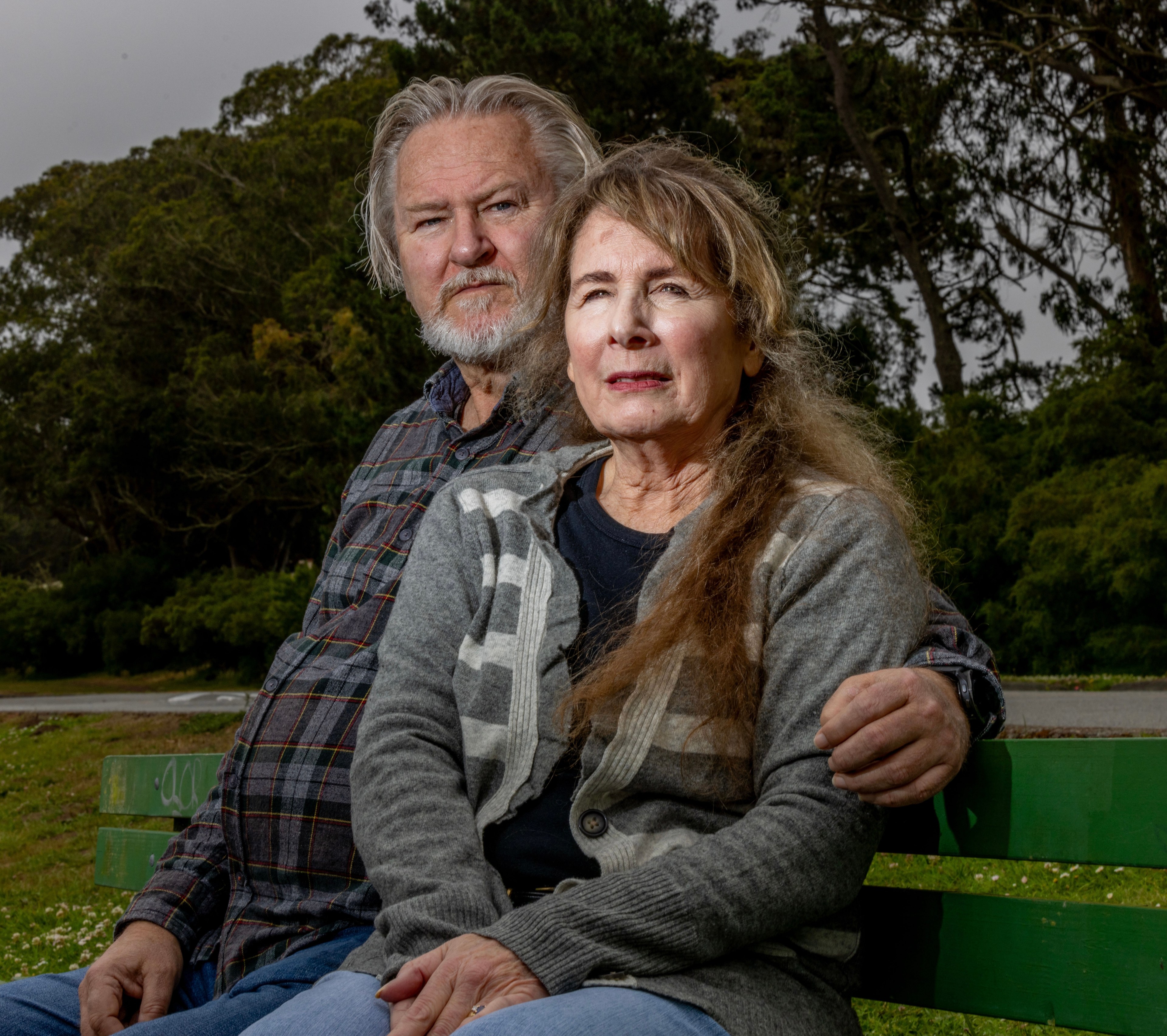 A mature couple sits closely on a green bench outdoors, surrounded by lush trees; the man has gray hair and a beard and wears a plaid shirt, while the woman has long hair and a gray cardigan.