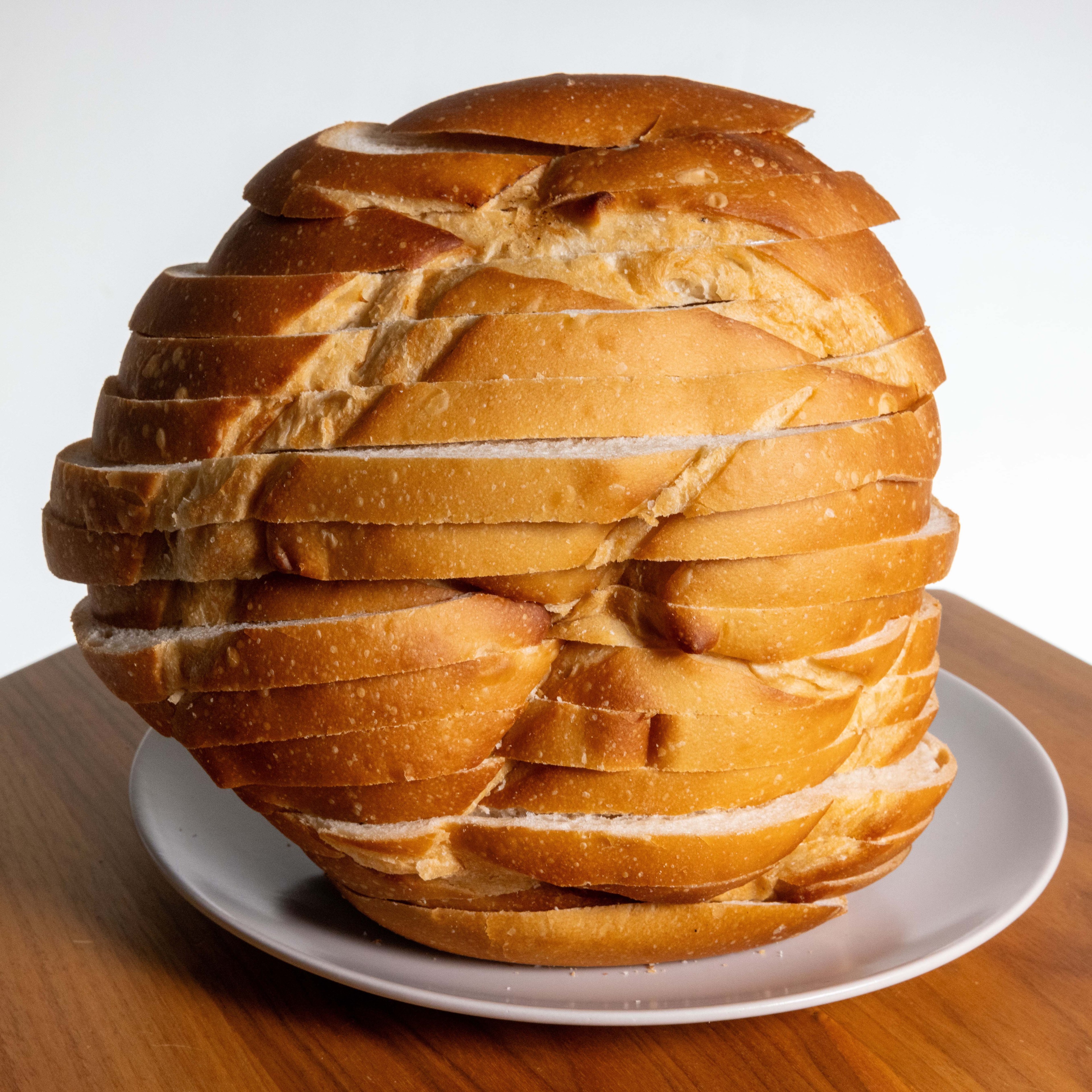 The image depicts a loaf of bread sliced and reassembled into a spherical shape, placed on a white plate on a wooden table, with a plain white background.