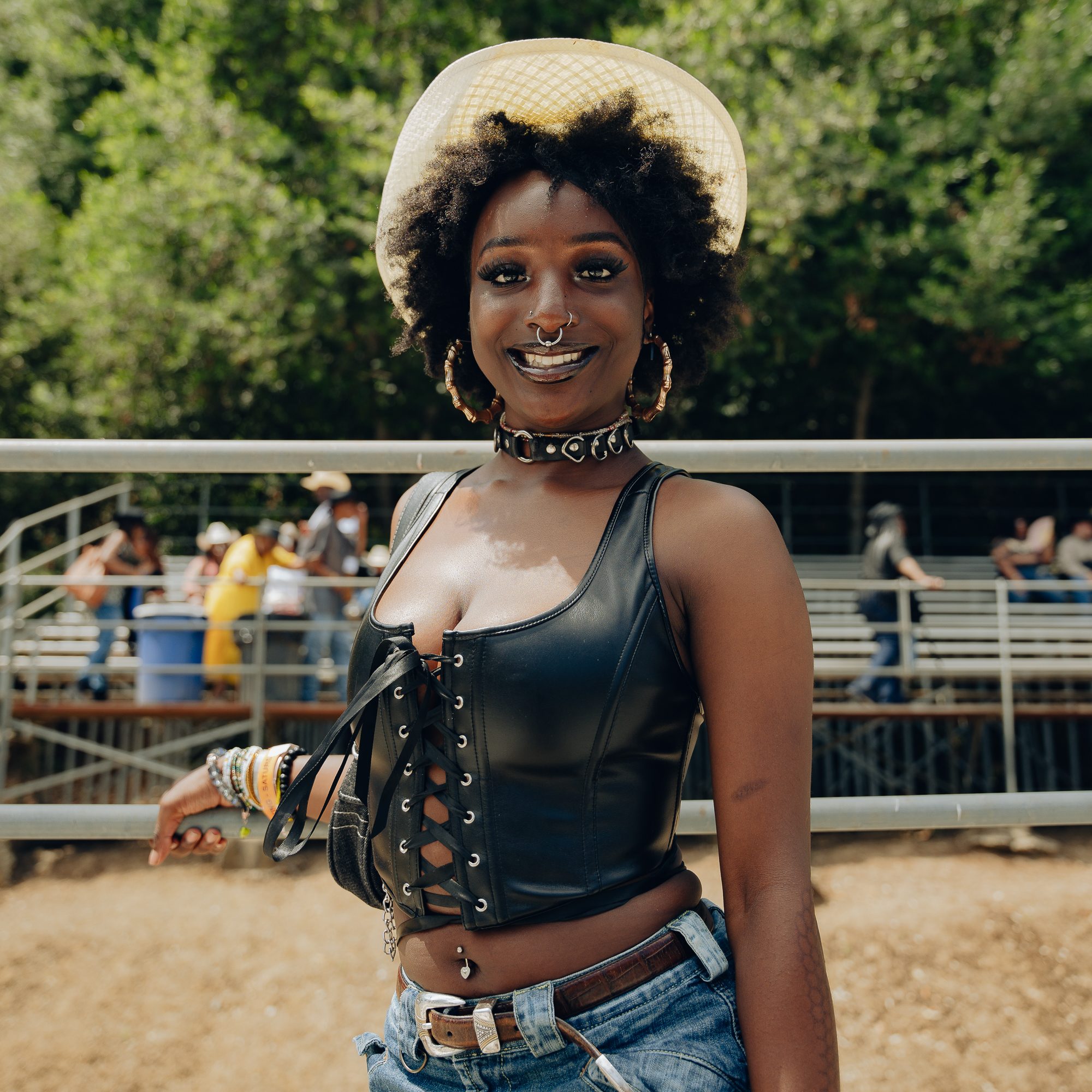 A smiling person wearing a cowboy hat, black leather lace-up top, and denim shorts stands outdoors against a fence, with a backdrop of trees and distant people.