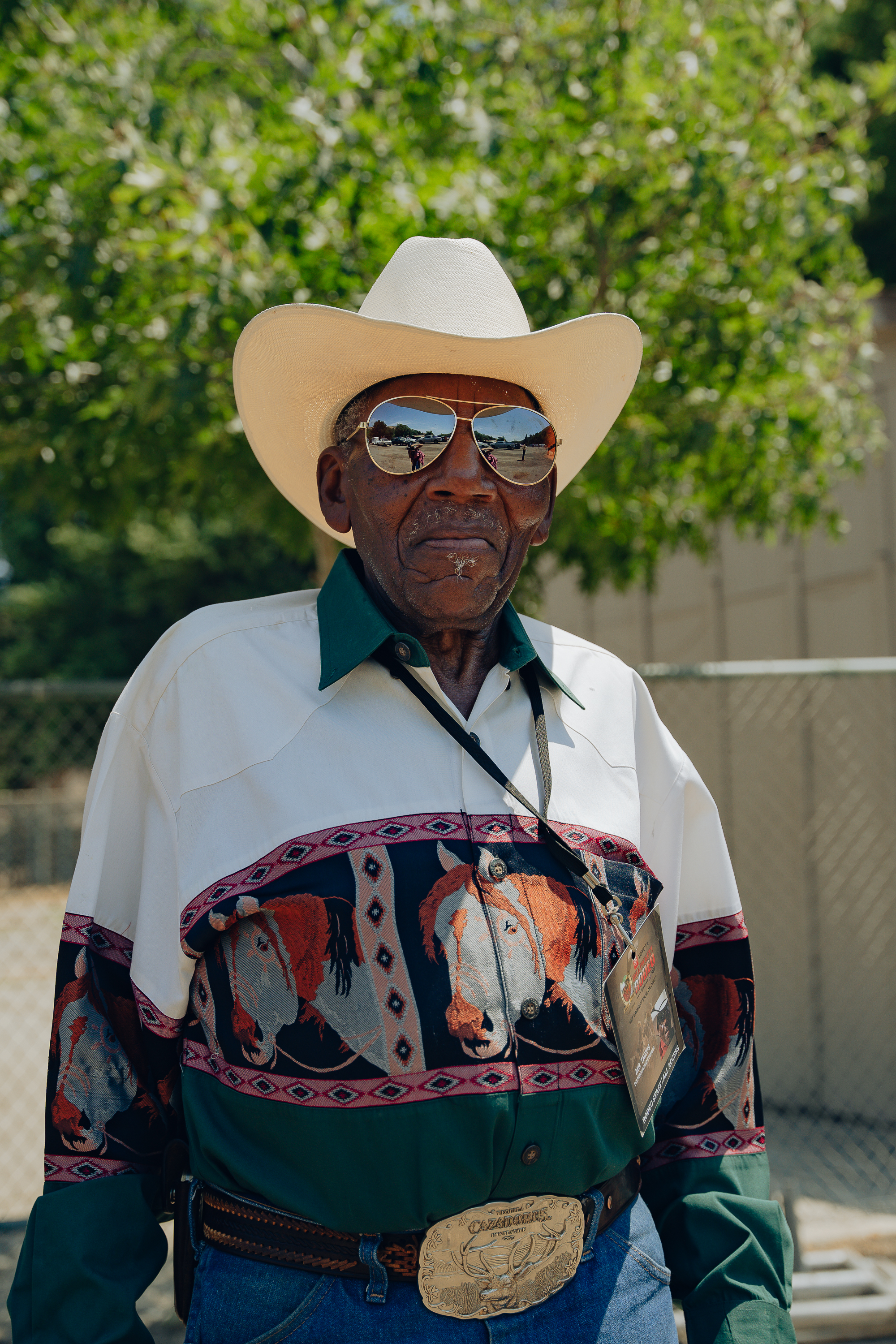 An elderly man wears a white cowboy hat, reflective sunglasses, and a colorful shirt with embroidered horses, paired with jeans and a large belt buckle. A tree is in the background.