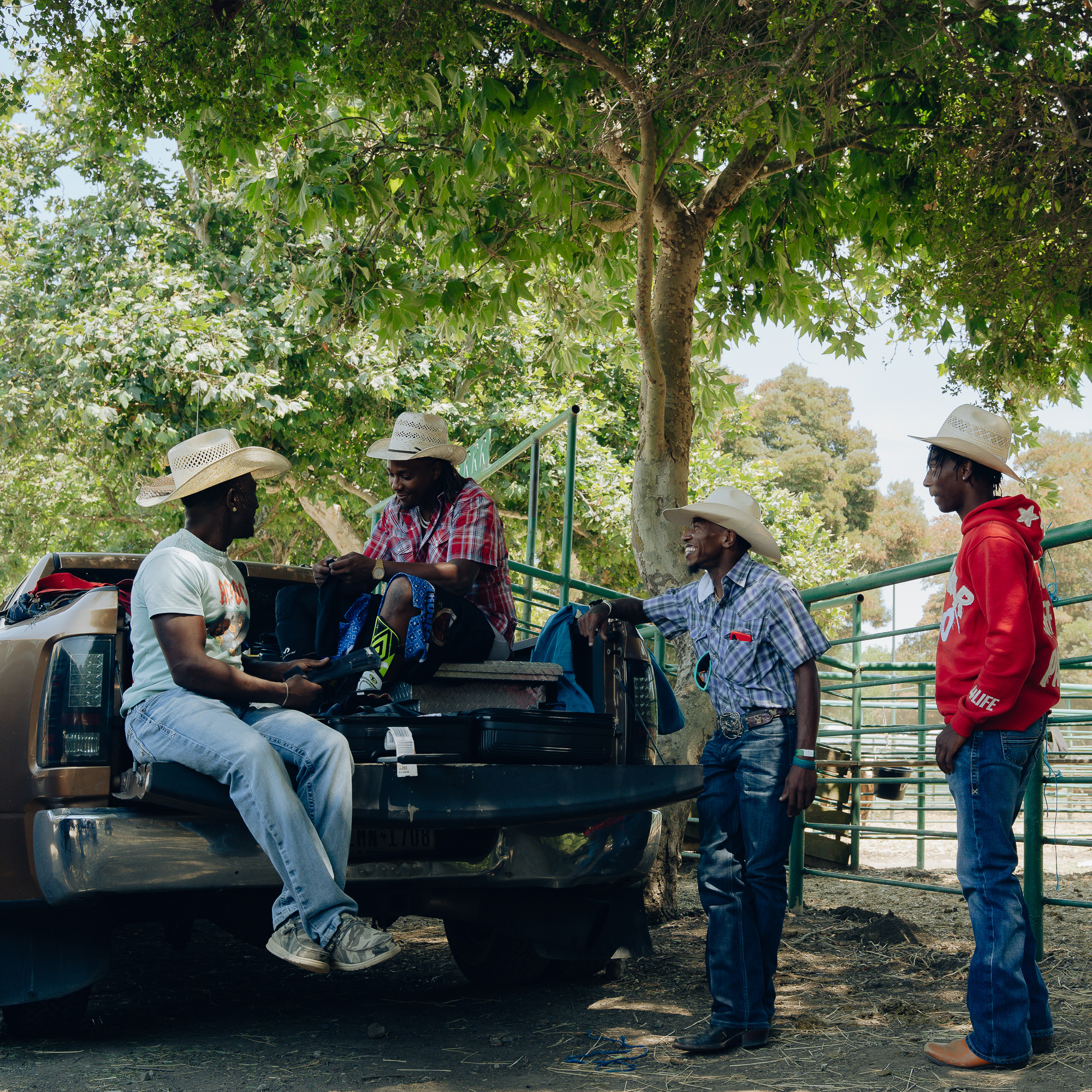 Four men wearing cowboy hats are gathered near a pickup truck parked under a tree. Two sit in the truck bed, while two stand beside it, smiling and chatting.