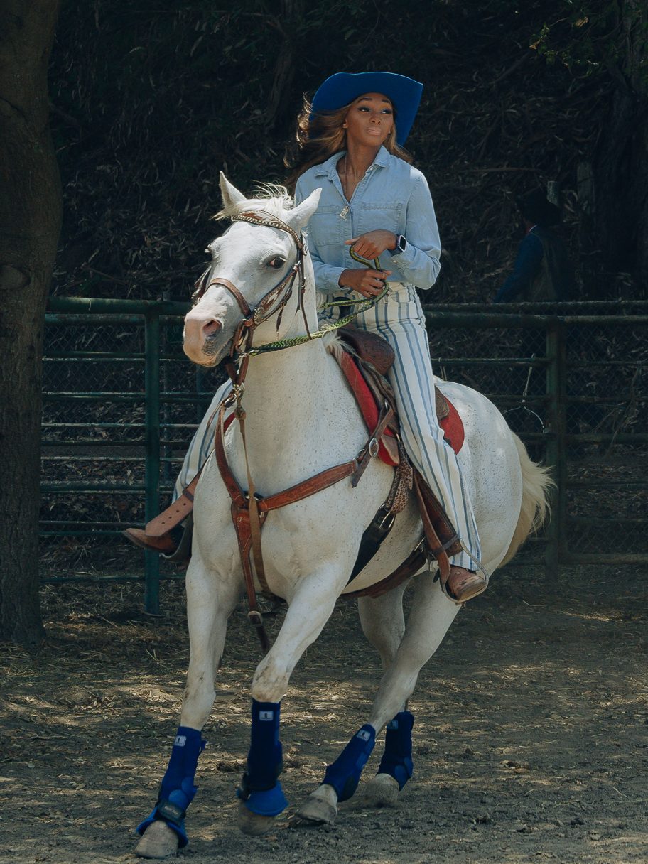 A person wearing a blue wide-brimmed hat and pinstriped pants rides a white horse with blue leg wraps in a fenced outdoor area, with people and trees in the background.