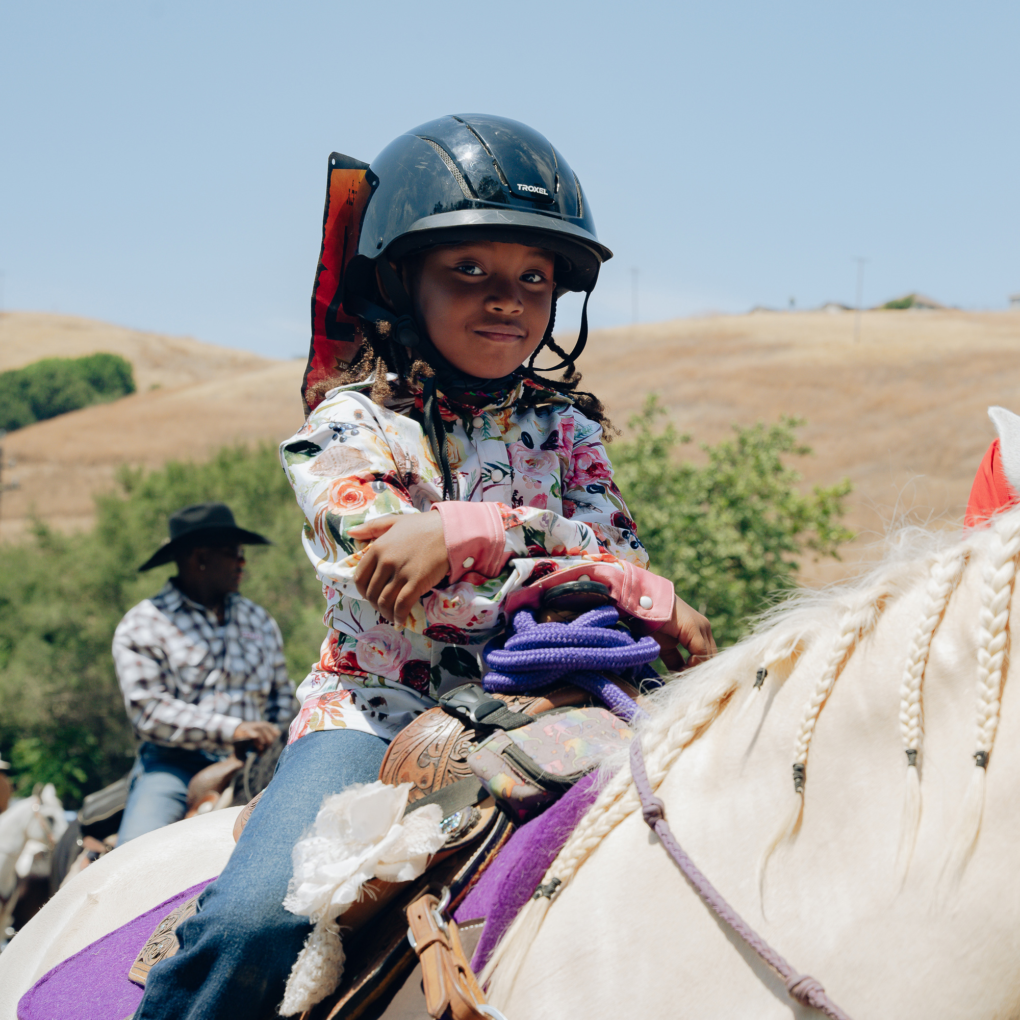 A child in a floral shirt and black helmet sits on a white horse with braids and a purple blanket. Behind, a person in a plaid shirt and cowboy hat is also riding.