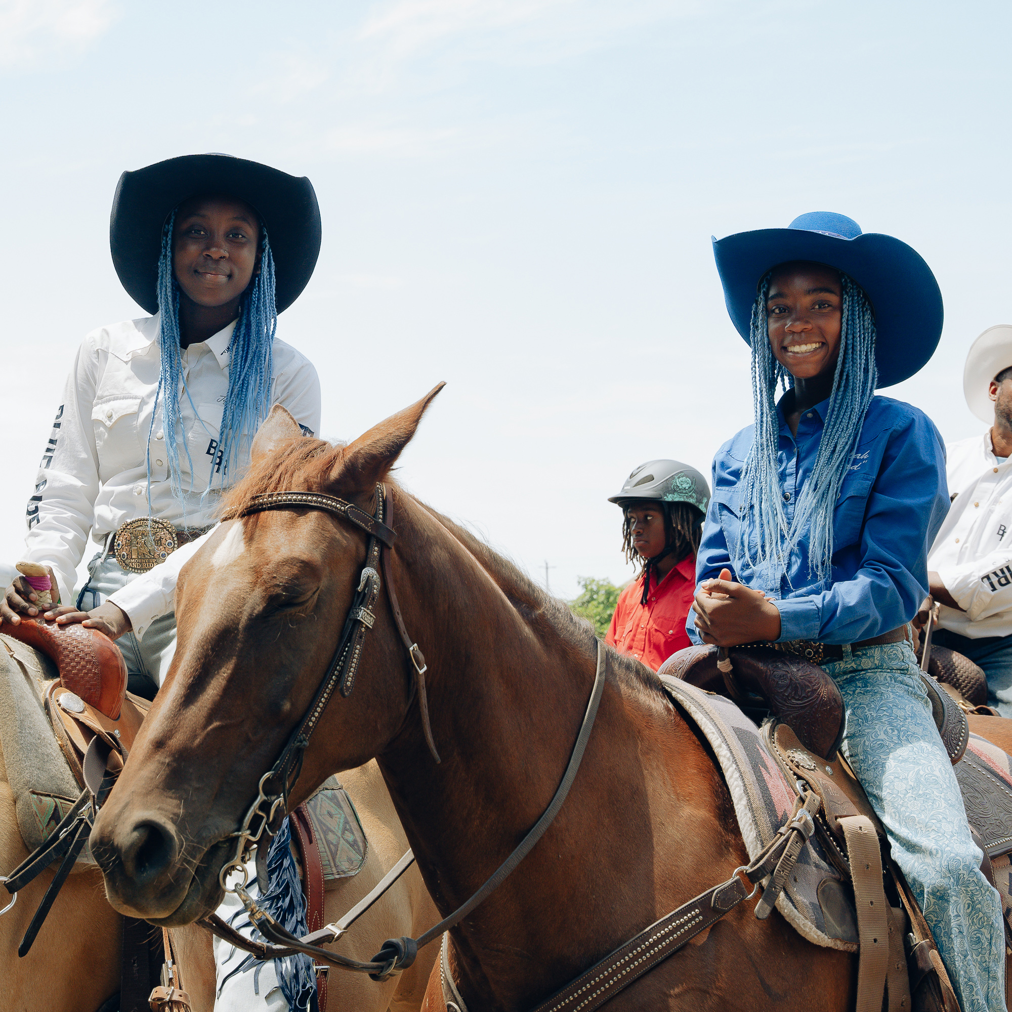 Two smiling individuals with blue braids and cowboy hats sit on horses, wearing western attire. The backdrop shows a few more riders and a partly cloudy blue sky.