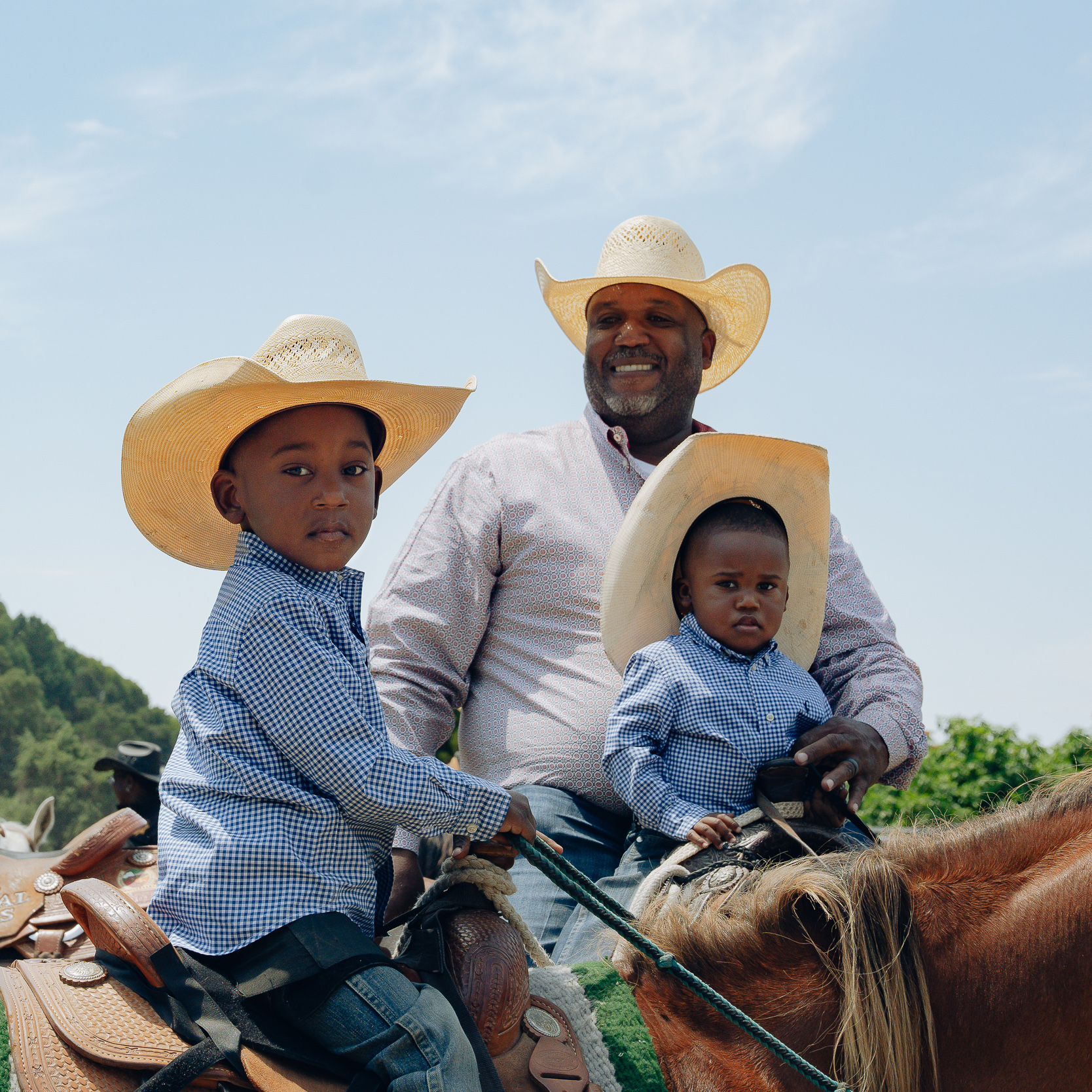 A smiling man and two young children, all wearing cowboy hats and checkered shirts, sit on horses with a clear sky and green trees in the background.